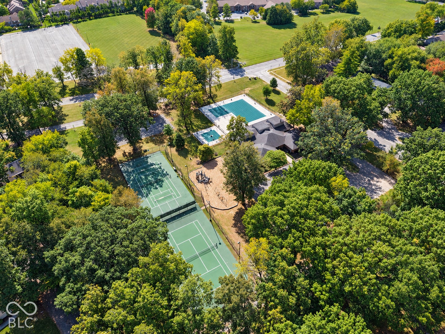 Aerial view showcasing an expansive residential compound featuring a resort-style swimming pool with integrated spa, professional-grade tennis court, manicured lawn, mature landscaping with established trees, and a modern residential structure set on substantial grounds surrounded by verdant acreage