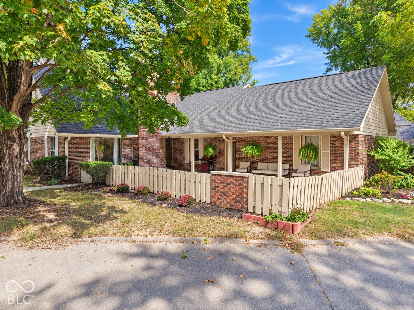 Charming brick ranch home featuring a wraparound covered porch with white railings and columns, expansive front lawn shaded by mature trees, and a gabled roofline with dimensional shingle detailing