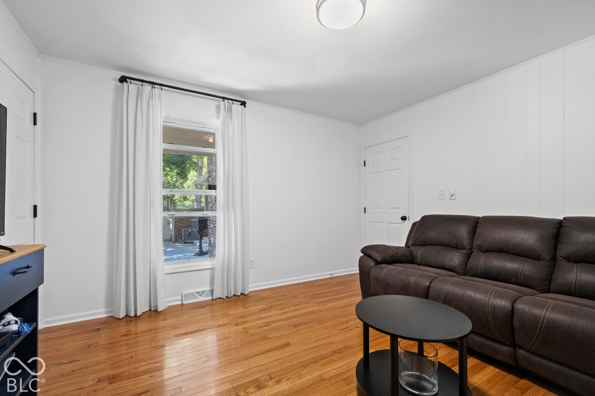 Bright living space with gleaming wood-finish flooring, floor-to-ceiling windows with sheer drapery, and recessed ceiling lighting