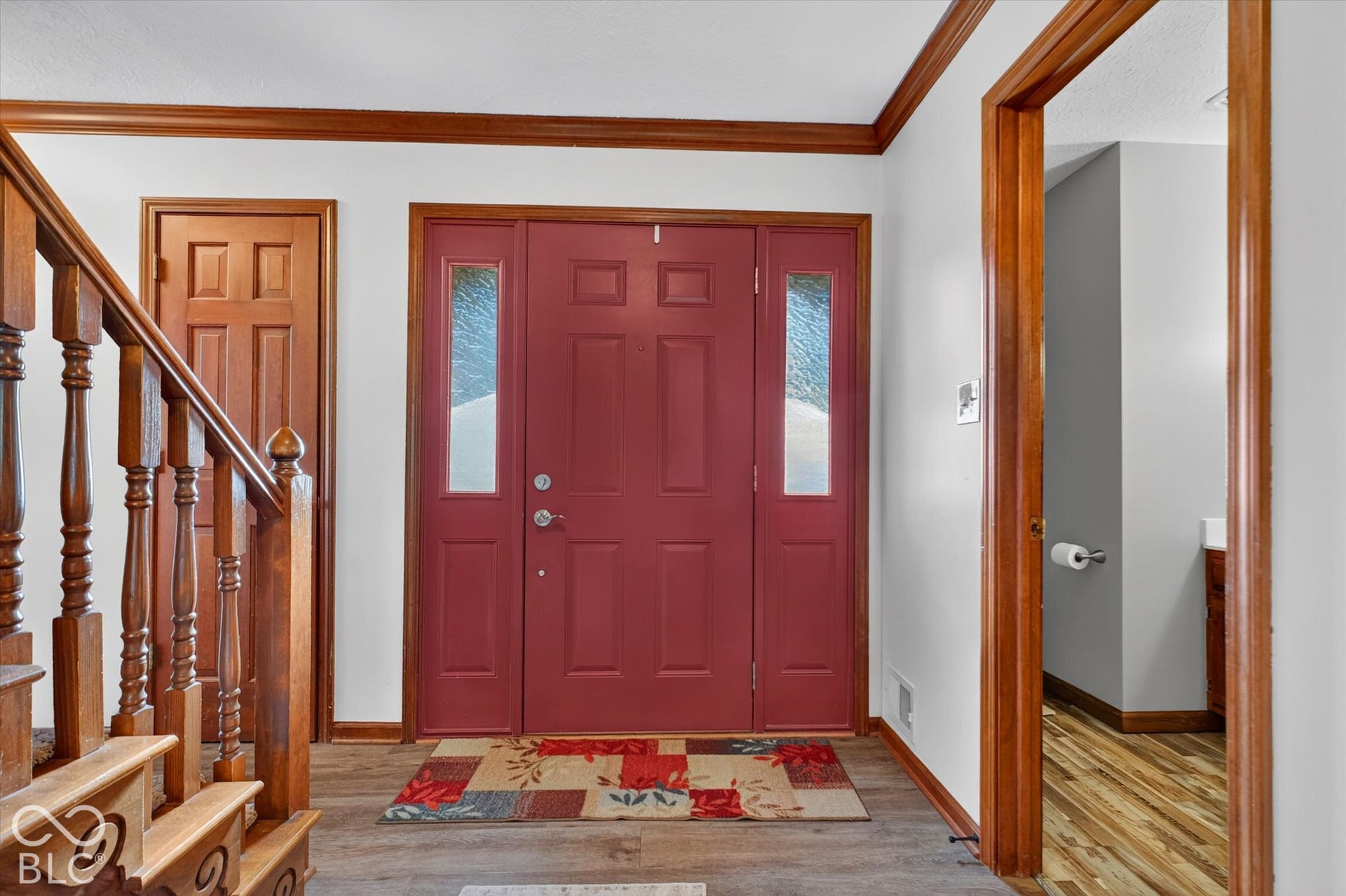 Welcoming foyer showcasing rich burgundy six-panel entry doors with sidelights, warm wood-finish trim and ceiling details, hardwood flooring, and open staircase with turned wooden balusters and newel posts