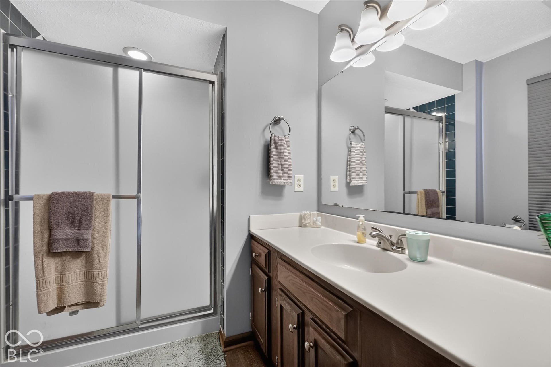 Shower with frosted glass enclosure, integrated lighting, wood-finish vanity with white countertop, and expansive wall mirror