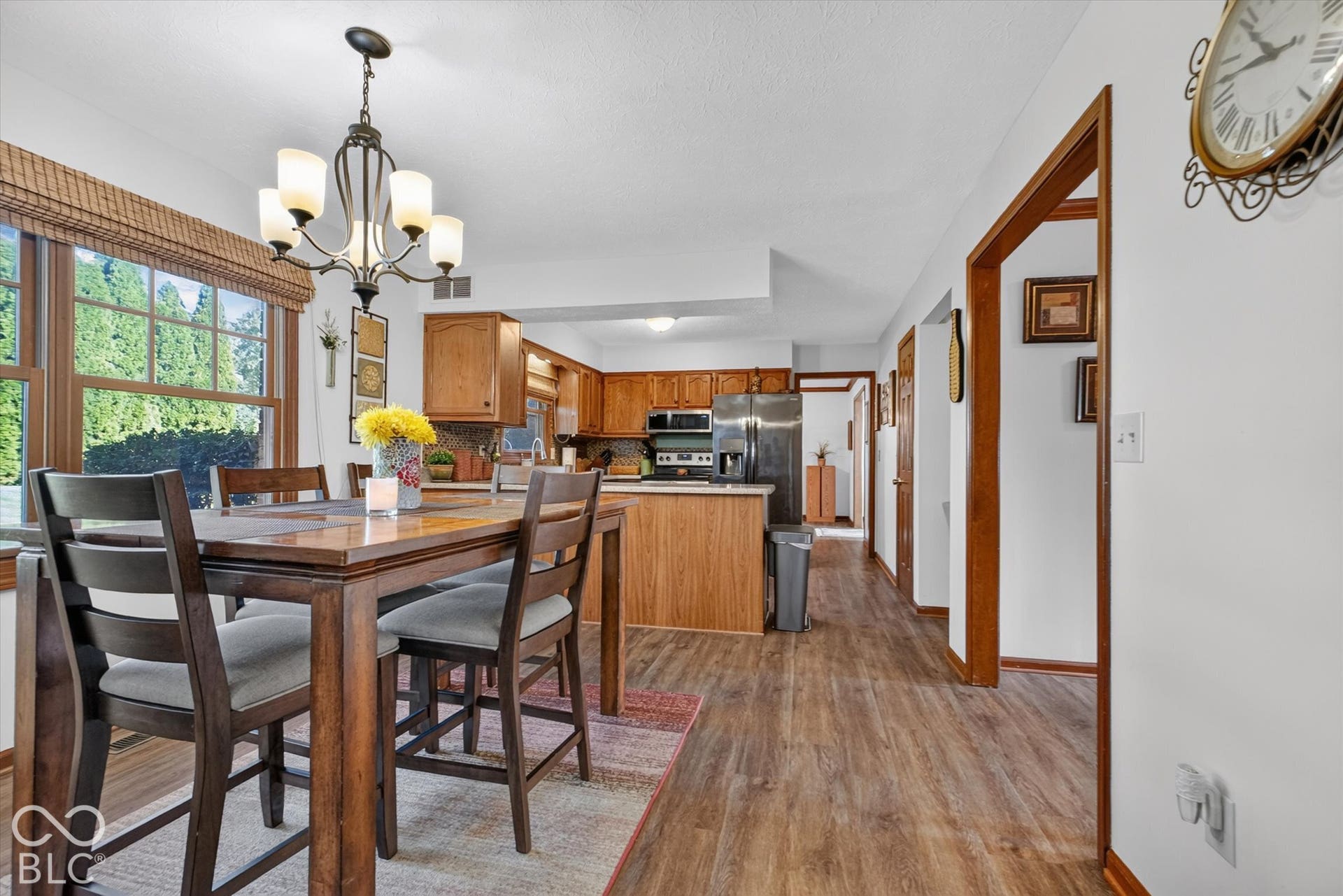 Dining area with a multi-light chandelier, large window with natural wood trim, and wood-finish flooring extending into the kitchen