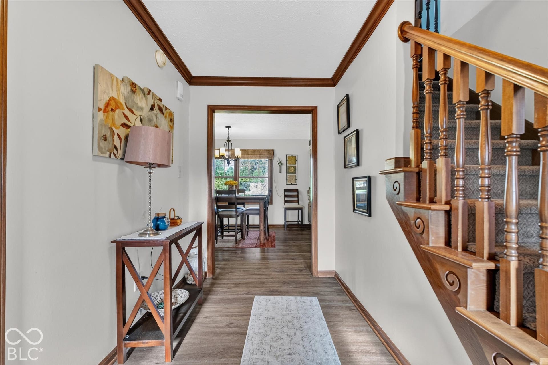 Wood-finish flooring, staircase with wood banister and balusters, wood trim and crown molding, and a framed doorway