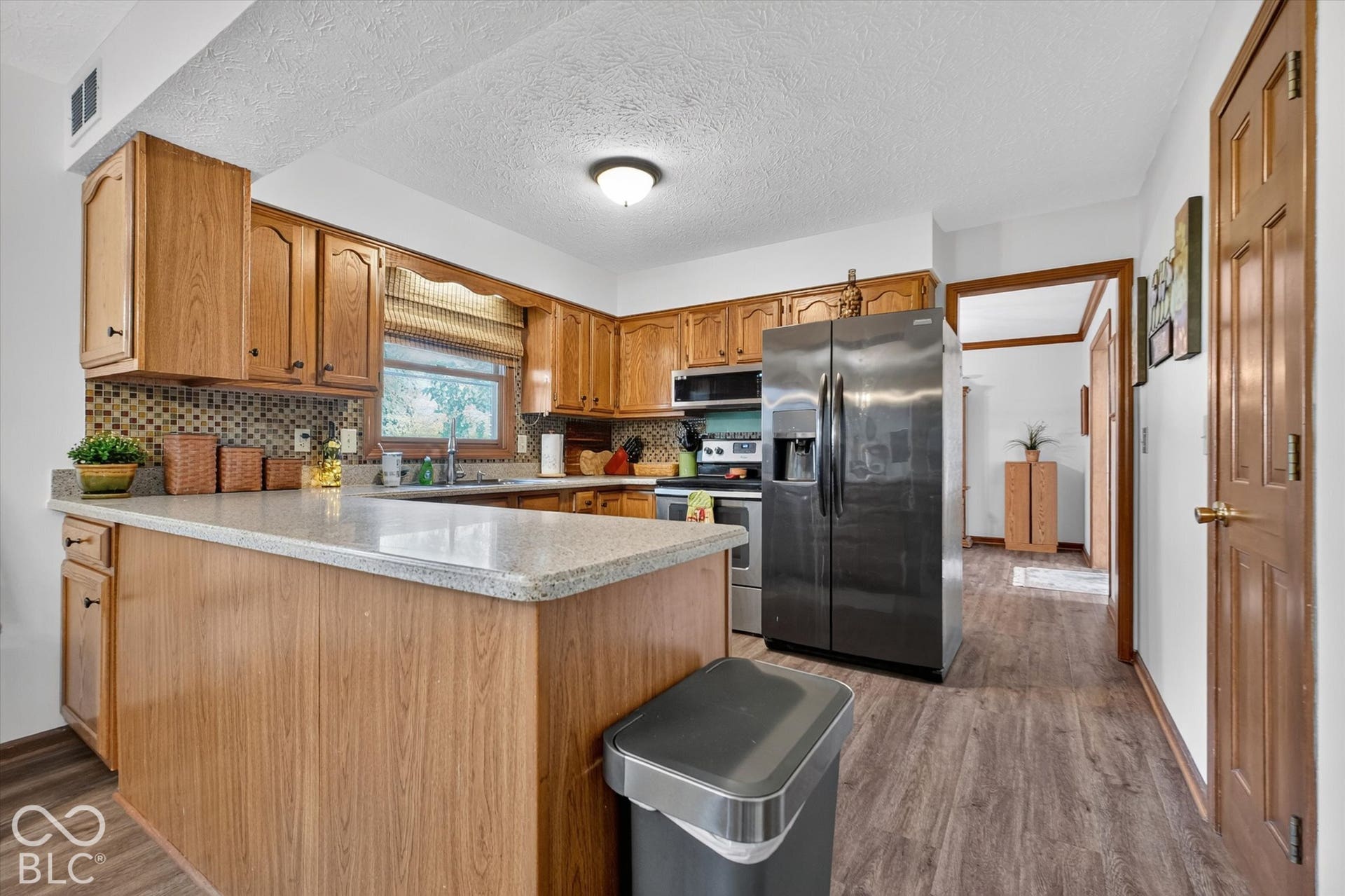 Wood-finish flooring, wood cabinetry, stone-look countertops, tile backsplash, and stainless steel appliances