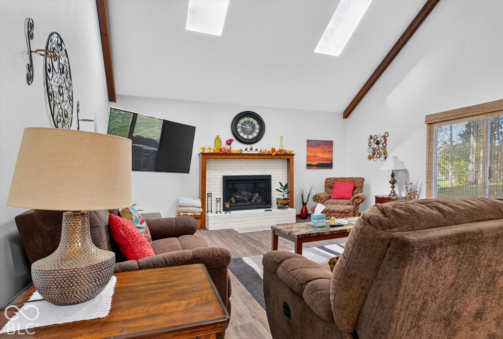 Vaulted ceiling with exposed wood beams, skylights, white brick fireplace, wood-finish flooring, and large windows