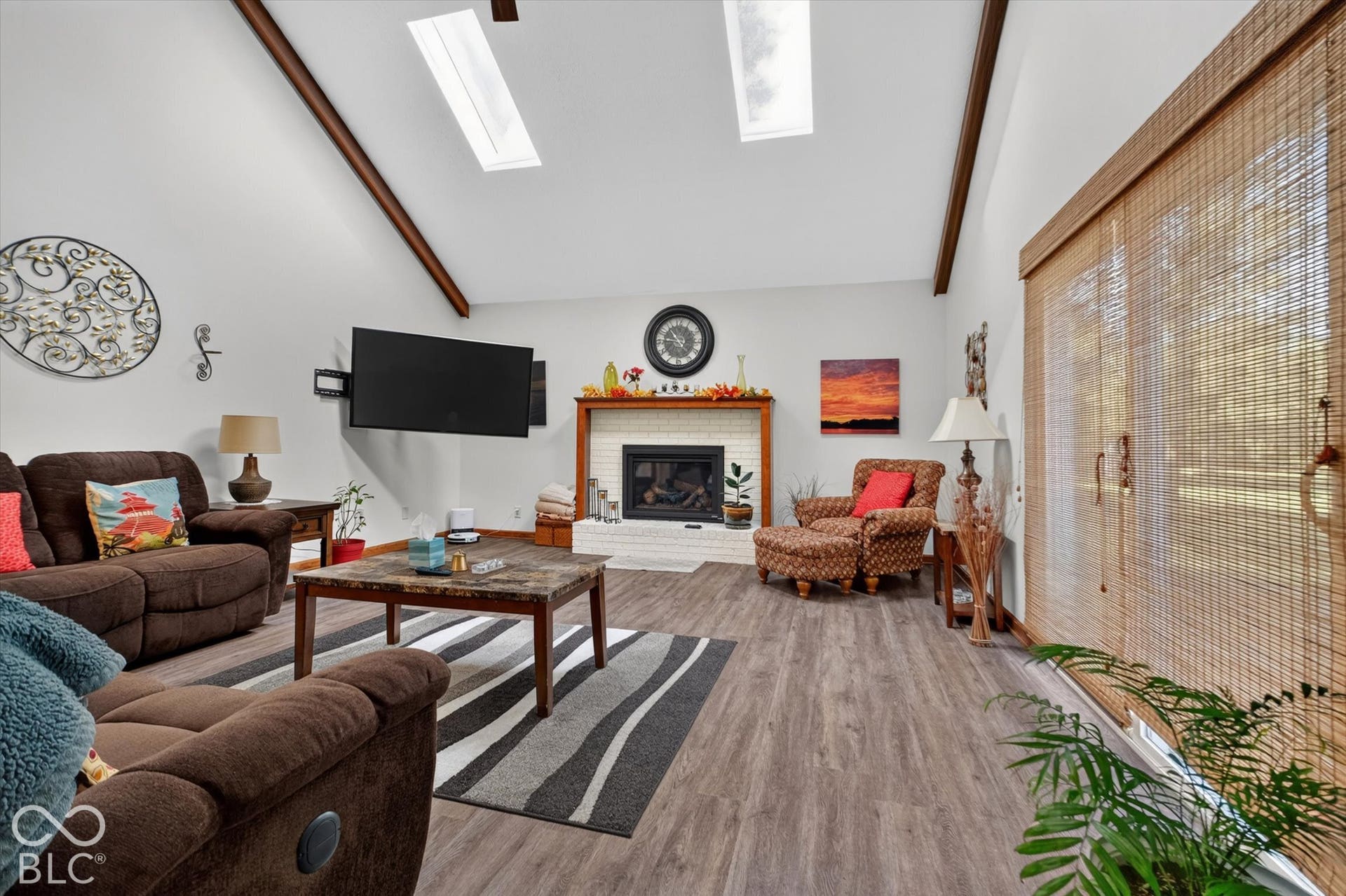 Vaulted ceiling with exposed beams, two skylights, wood-finish flooring, white brick fireplace with mantel, and sliding glass door
