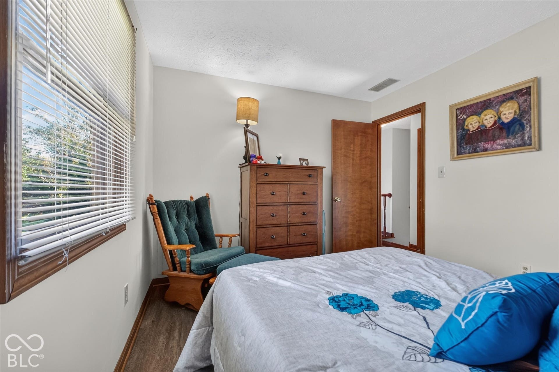 Comfortable secondary bedroom with wood-finish flooring, large window with horizontal blinds providing natural light, wooden dresser with integrated lamp, and pendant lighting fixture