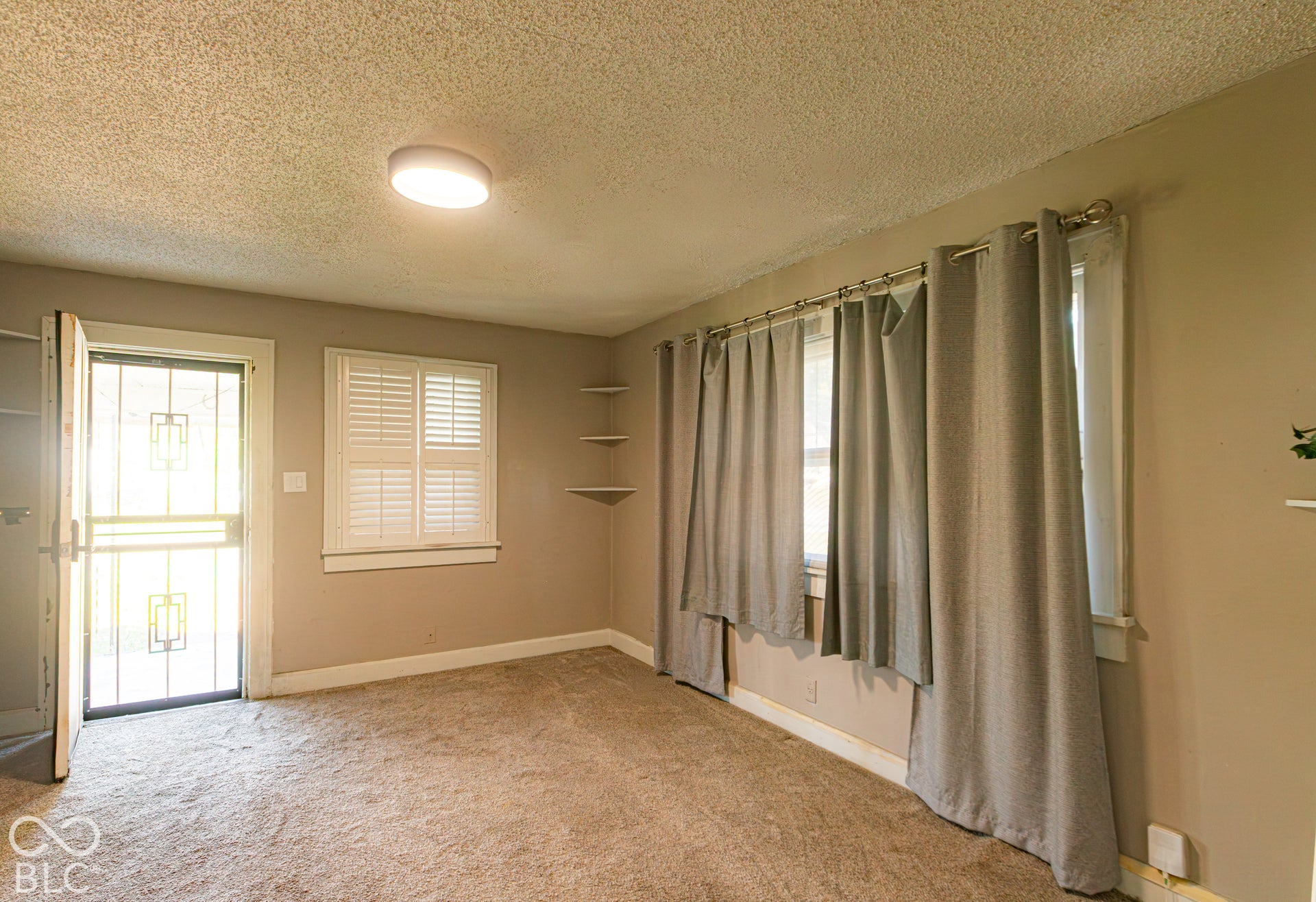 Neutral wall paint, ceiling-mounted light fixture, white plantation shutters, built-in corner shelving, and carpet flooring