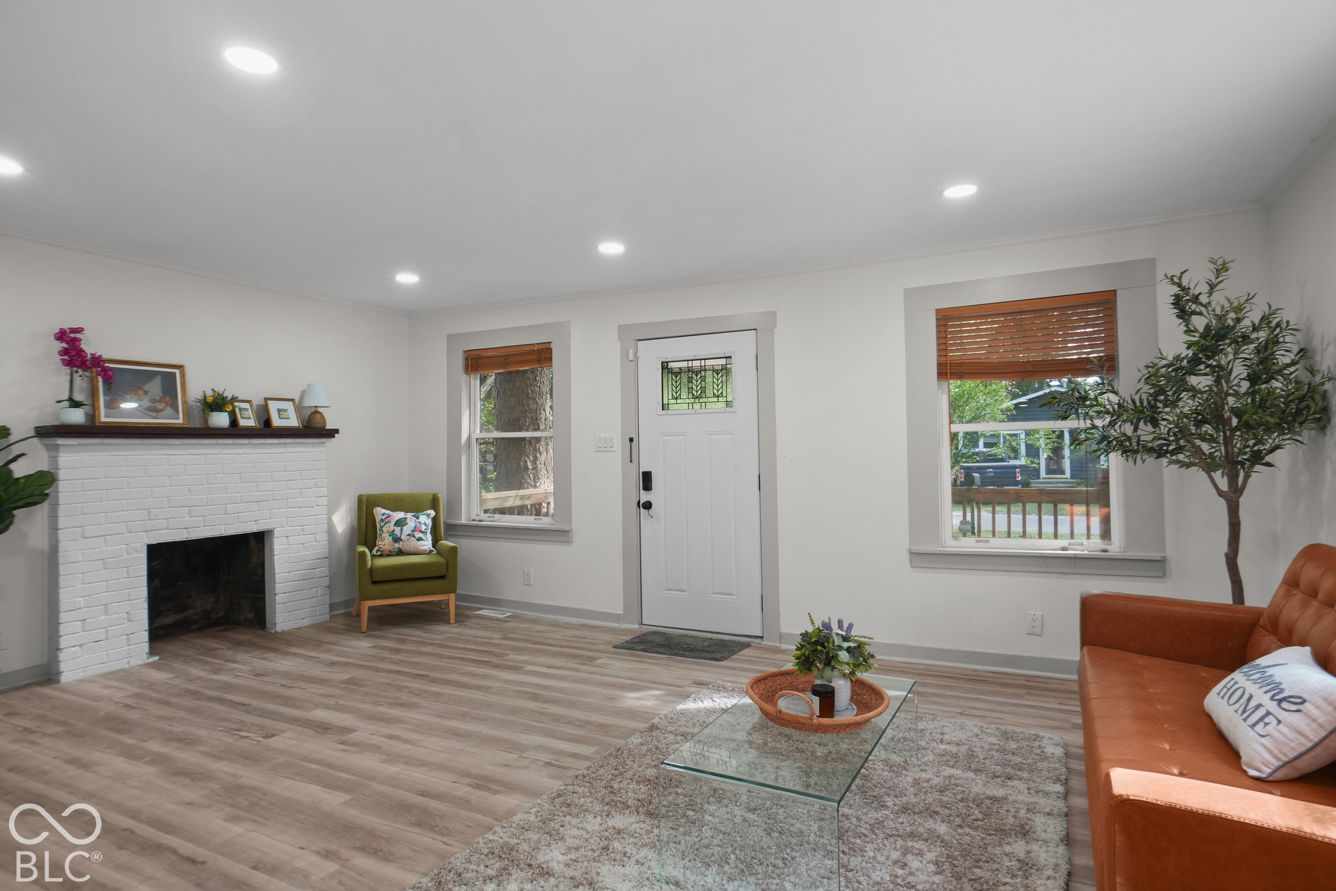 Recessed lighting, wood-finish flooring, white brick fireplace, white entry door with decorative glass, and window with wood blinds