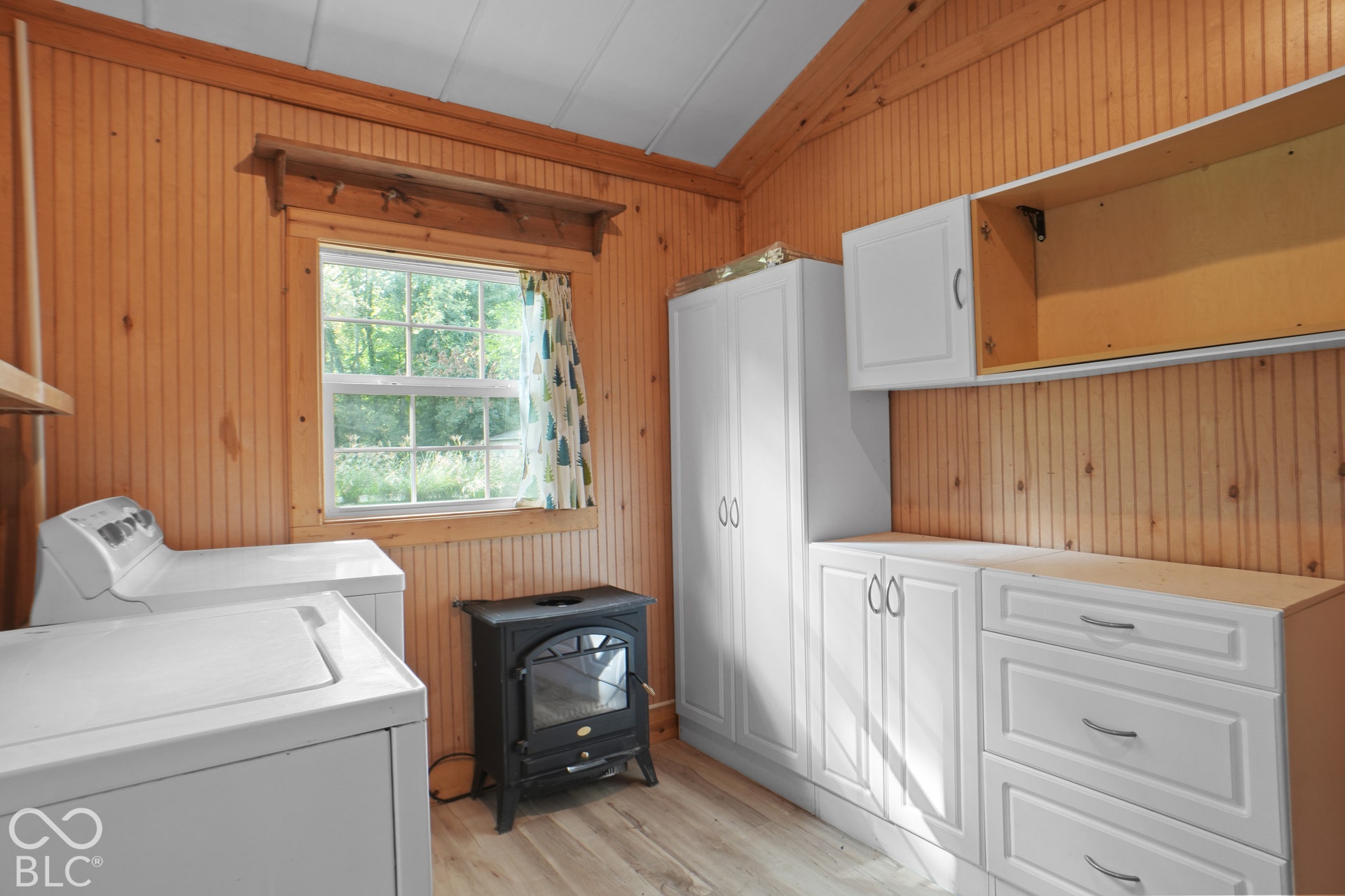 Wood-paneled walls and ceiling, wood-finish flooring, white cabinetry with silver hardware, and a black cast-iron stove