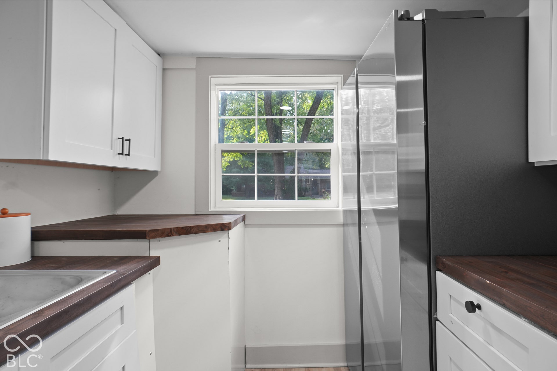 White shaker cabinetry with matte black hardware, wood-finish countertops, stainless steel refrigerator, and a double-hung window