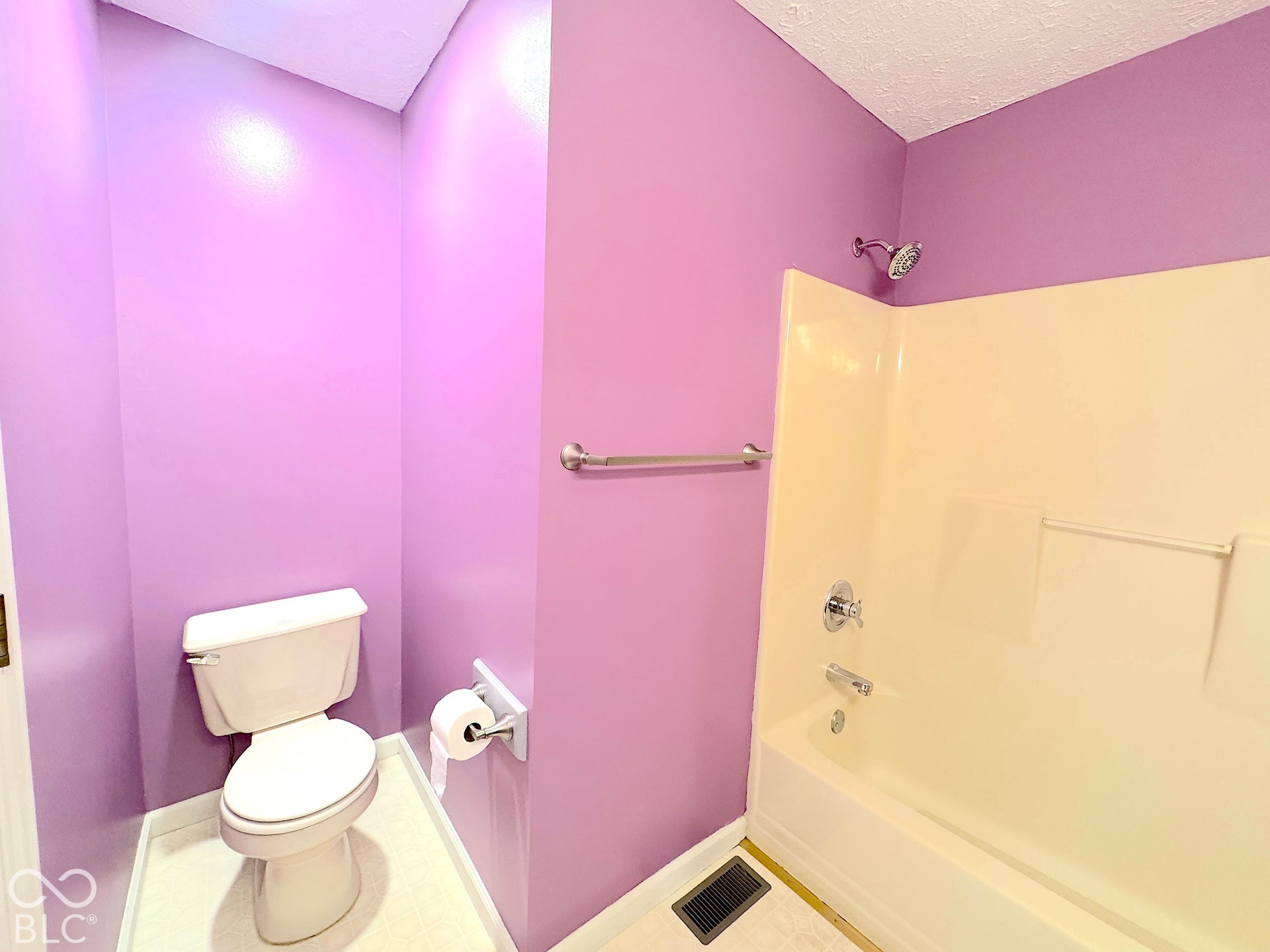 White toilet with a wall-mounted paper holder, chrome towel bar, and a white tub/shower combination with chrome fixtures