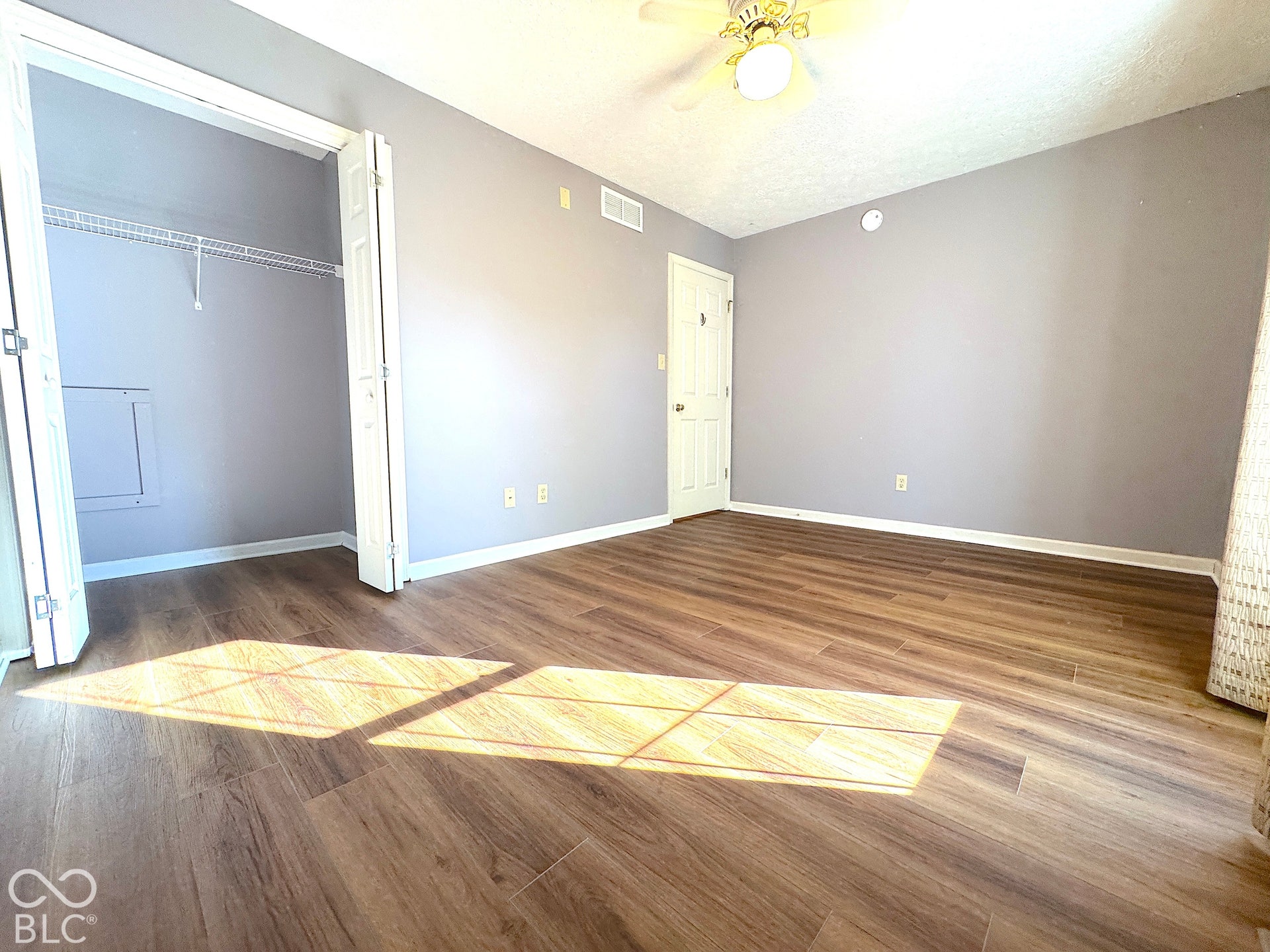 Wood-finish flooring, ceiling fan with light fixture, light gray wall paint, white trim, and bi-fold closet doors