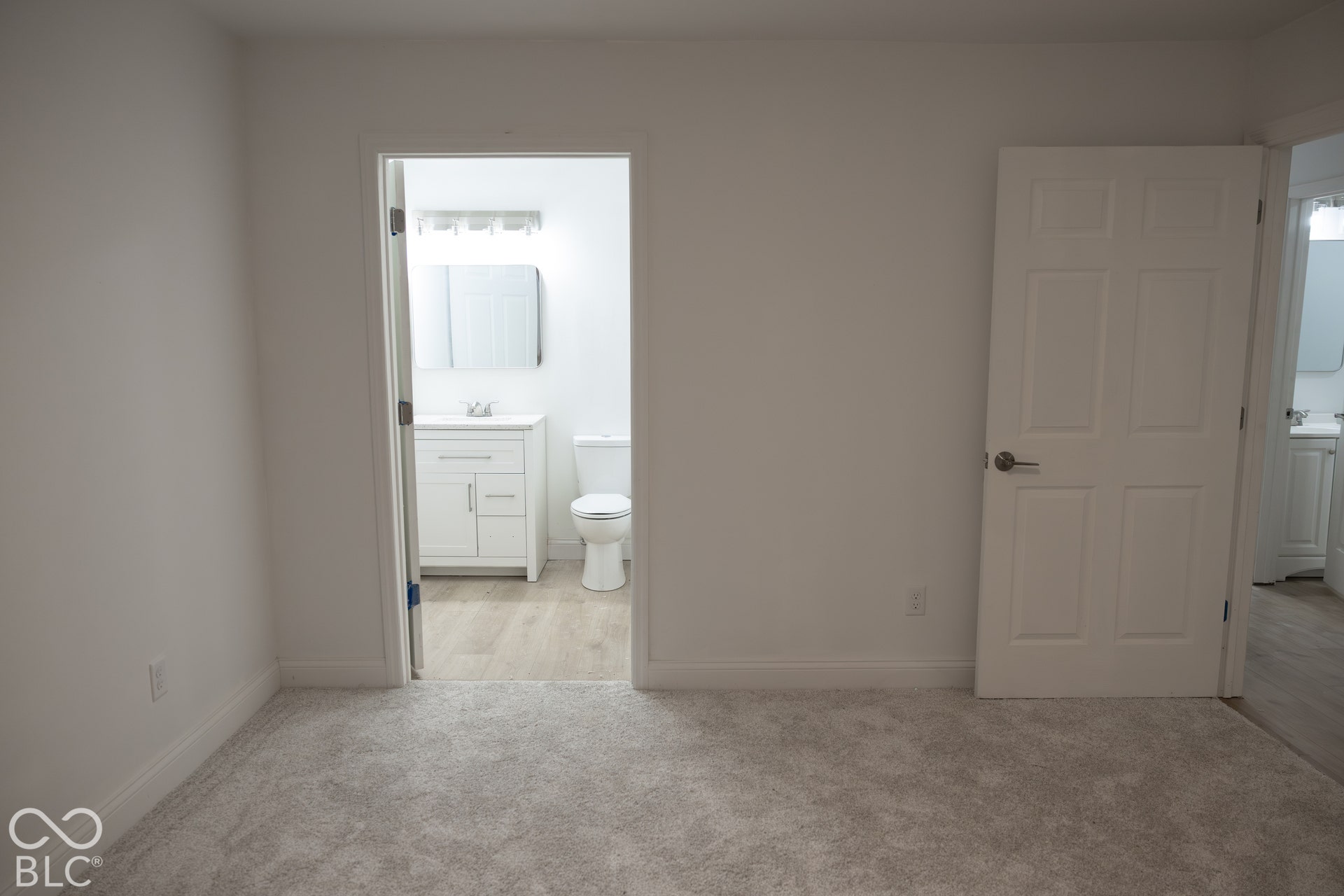Plush carpet flooring, white interior doors with panel detailing, white walls, and a bathroom featuring a white vanity with integrated sink and a wall-mounted light fixture