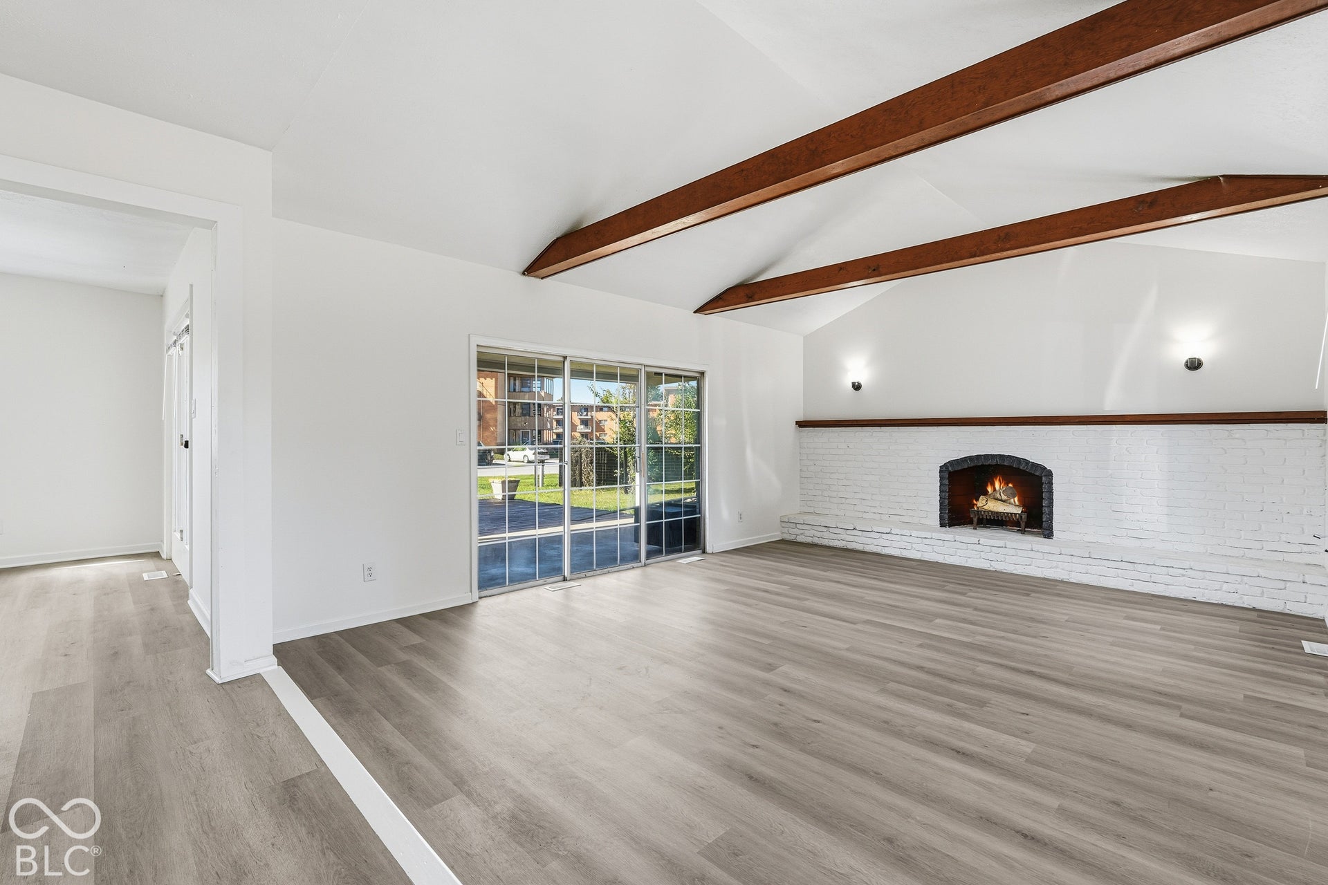 Vaulted ceiling with exposed wood beams, wood-finish flooring, white painted brick fireplace, and sliding glass doors to the exterior