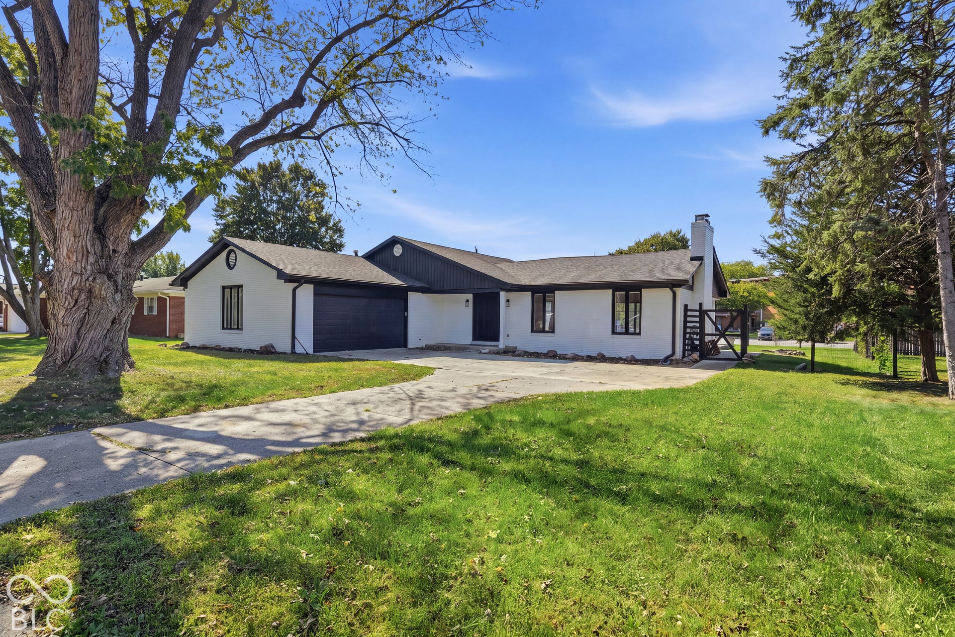 Exterior featuring a white facade, black trim windows, an attached garage, a paved driveway, and mature landscaping