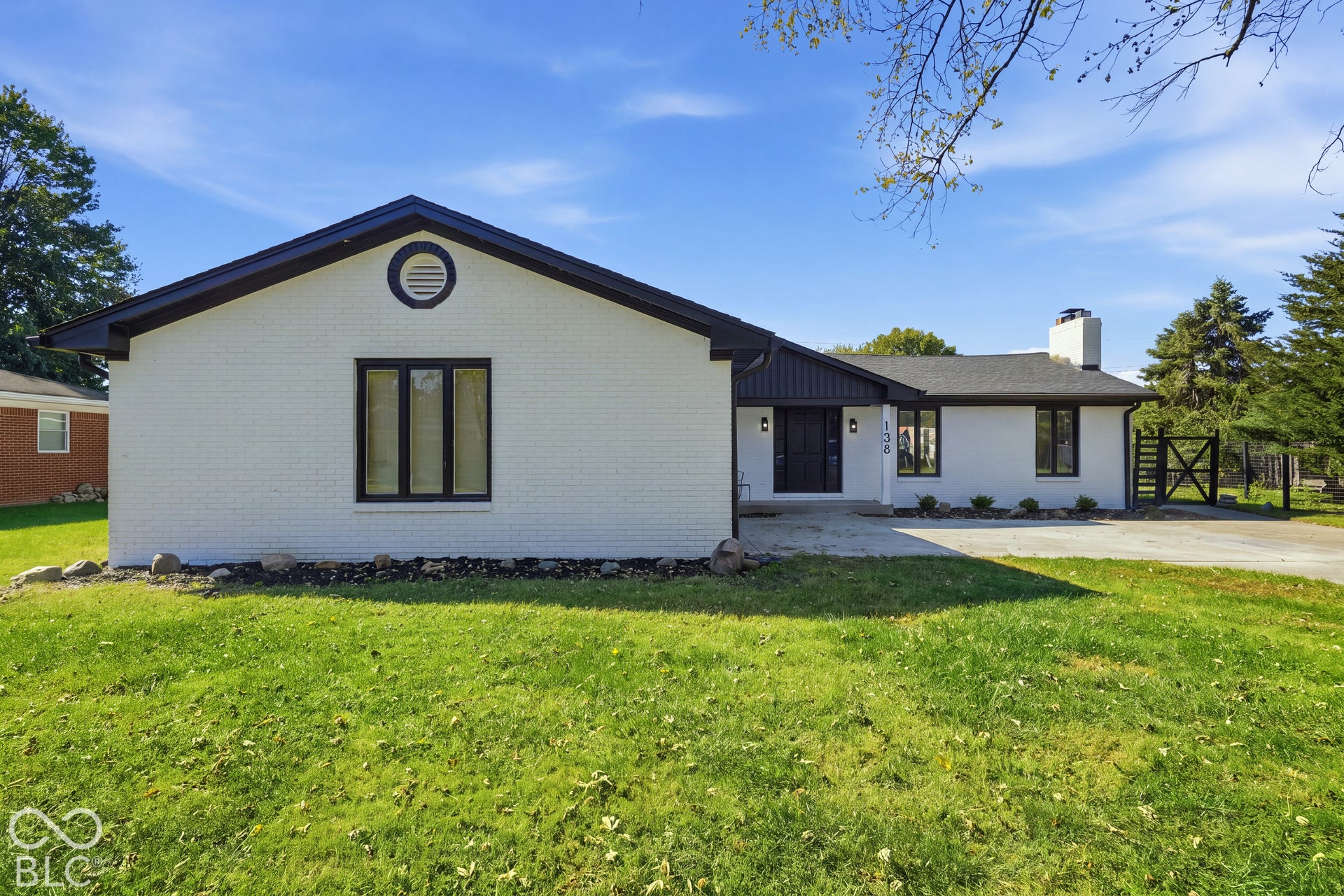 White brick exterior with black trim, dark shingle roof, attached chimney, and a gravel driveway