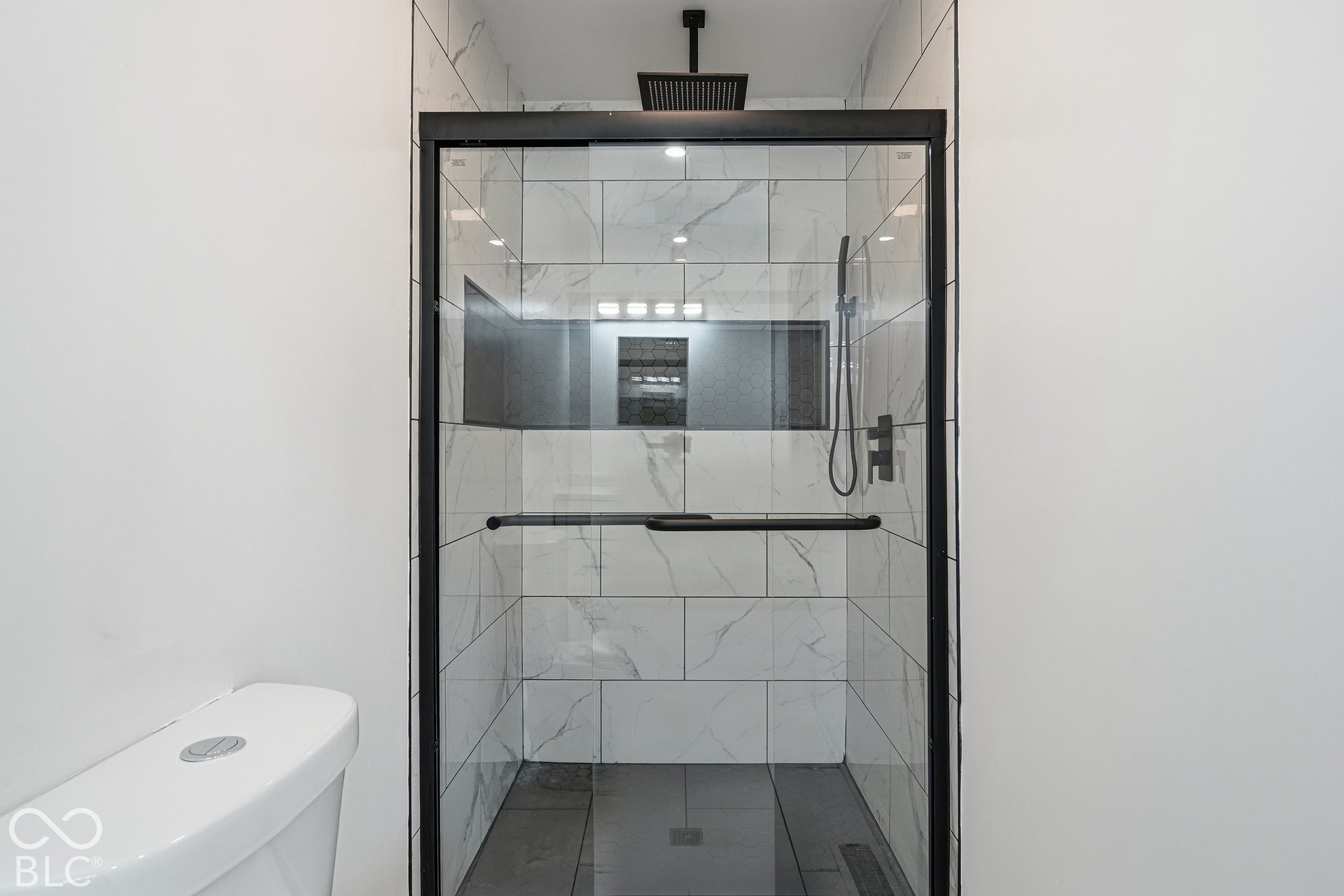 Glass-enclosed shower with a matte black frame, rain shower head, hand-held shower fixture, built-in recessed shelf, and white tile surround