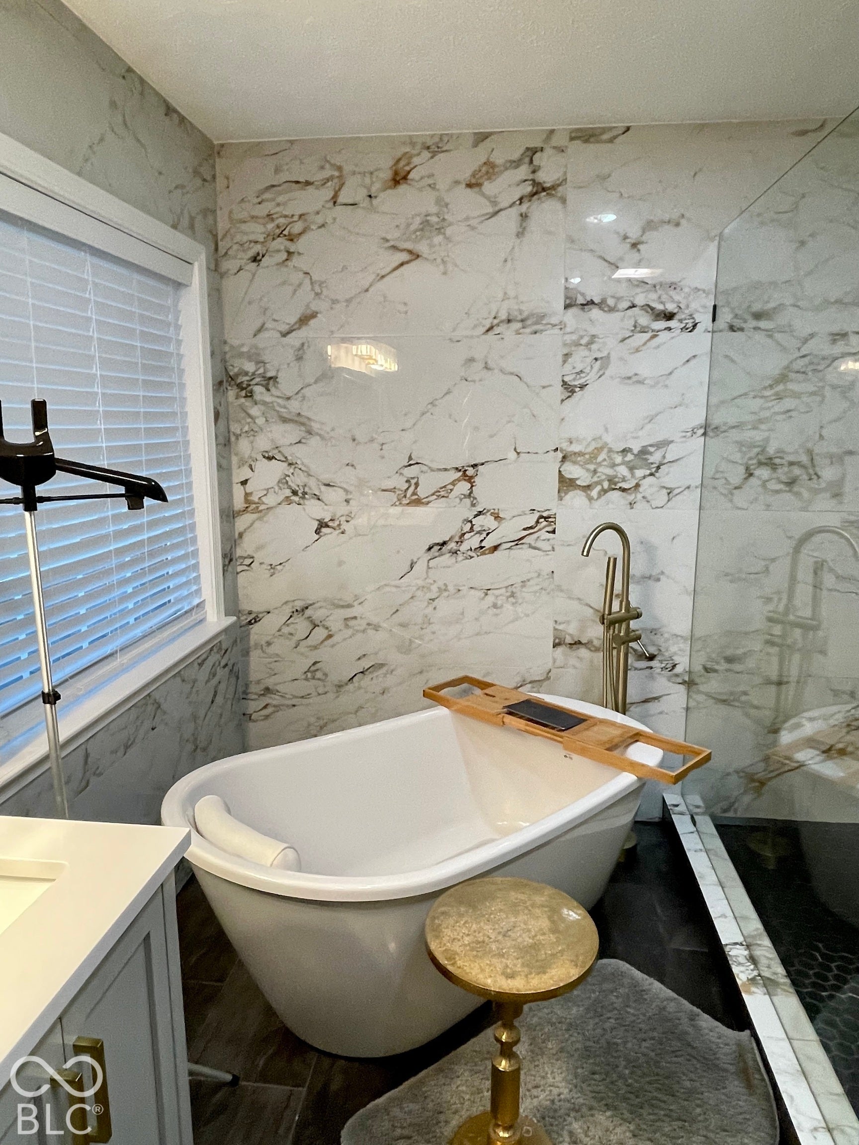 Freestanding soaking tub with a floor-mounted gold-tone faucet, complemented by large format marbled tile walls and dark wood-finish flooring