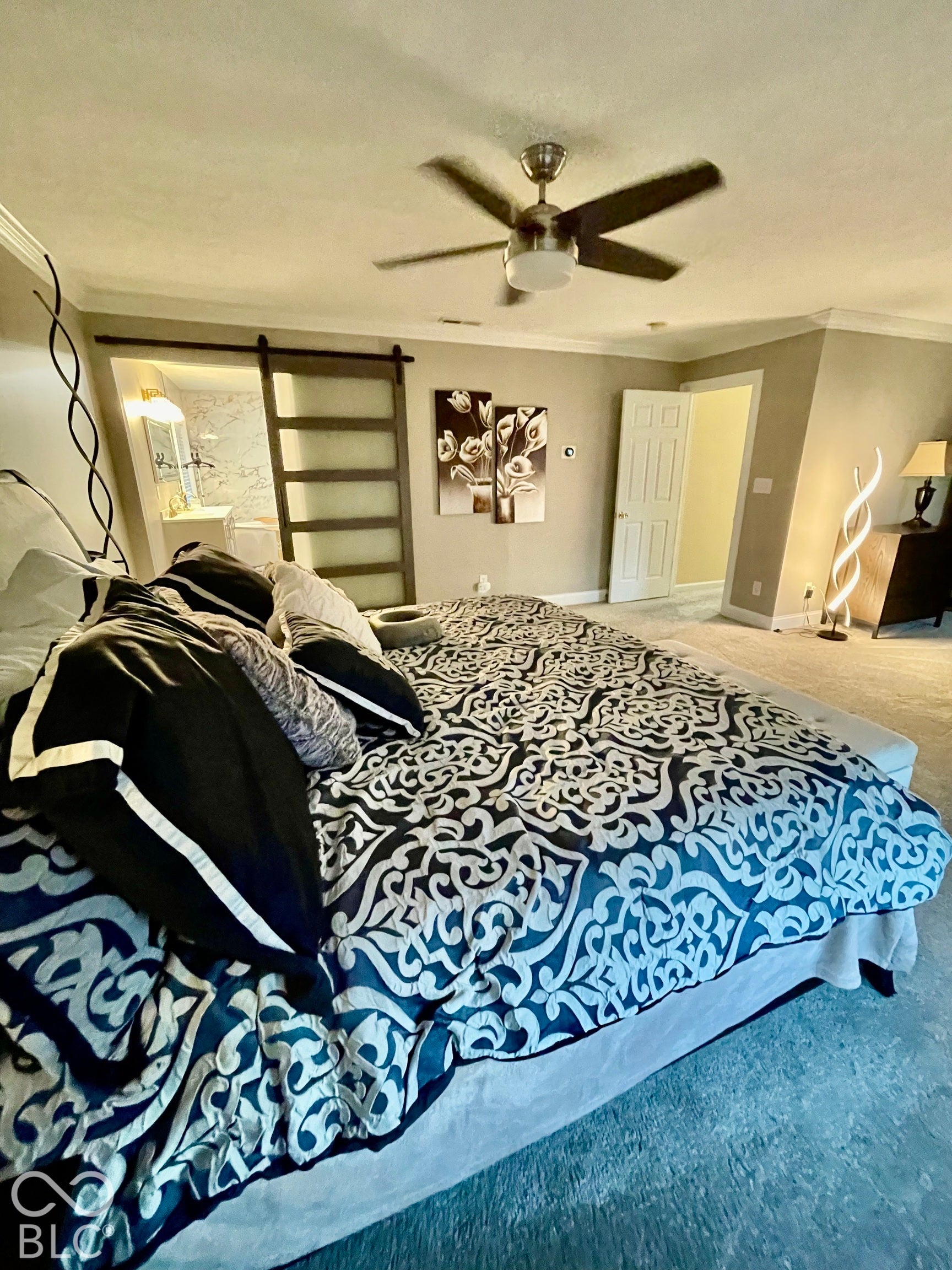 Modern ceiling fan with light fixture, carpeted flooring, barn door entry to bathroom, and white bathroom vanity with double sinks
