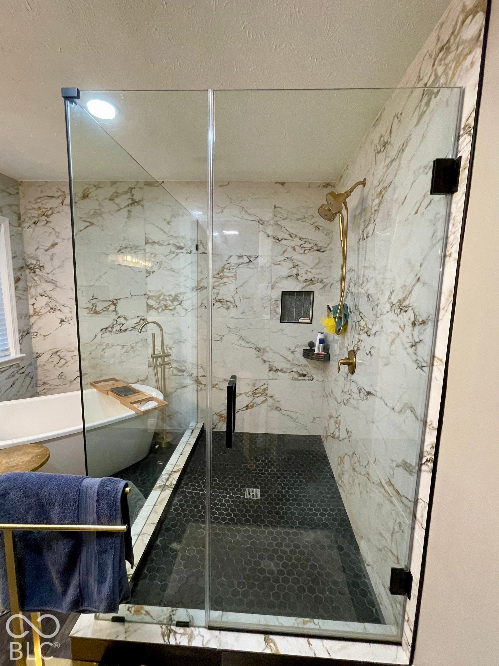 Spacious walk-in shower with clear glass enclosure, white and gold veined tile surround, black hexagonal floor tile, built-in shower niche, and a freestanding soaking tub