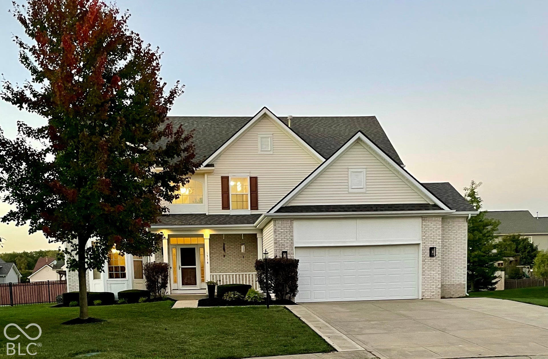 Light-colored siding exterior with a dark shingled roof, a two-car garage, and a covered front porch featuring white railings and dark sconces