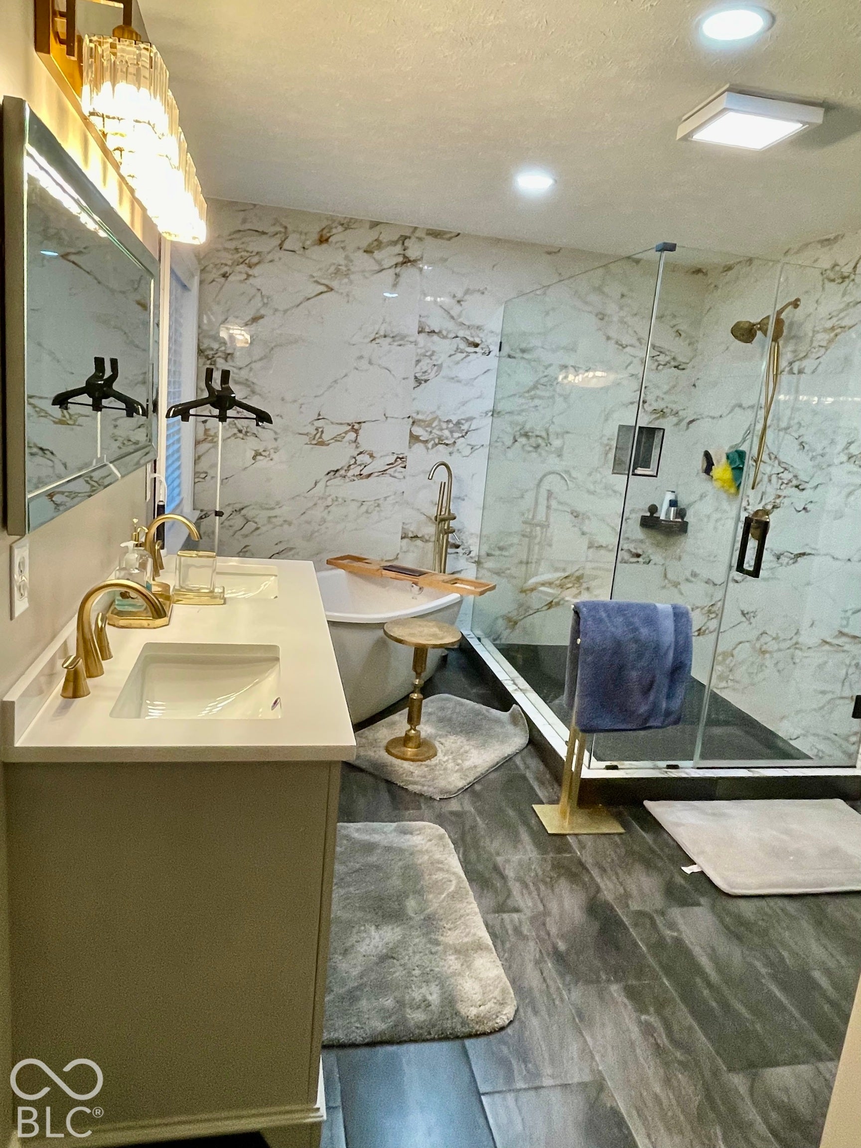 Dual vanity with undermount sinks and gold-finish faucets, freestanding bathtub, glass-enclosed shower, wall-mounted tile, and wood-finish flooring