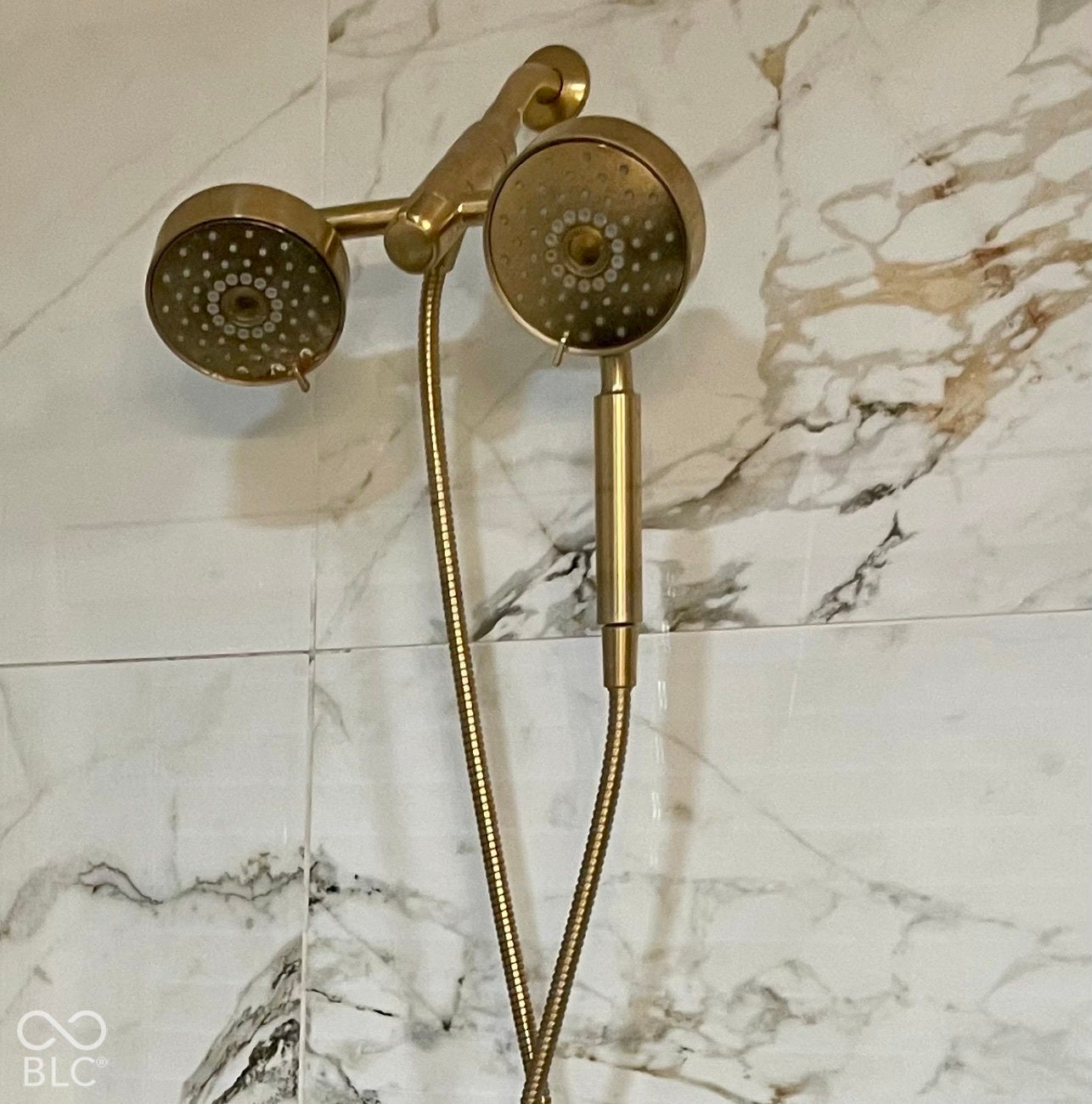 Dual shower heads with a brass finish and a flexible hose, against white and grey veined tile surround