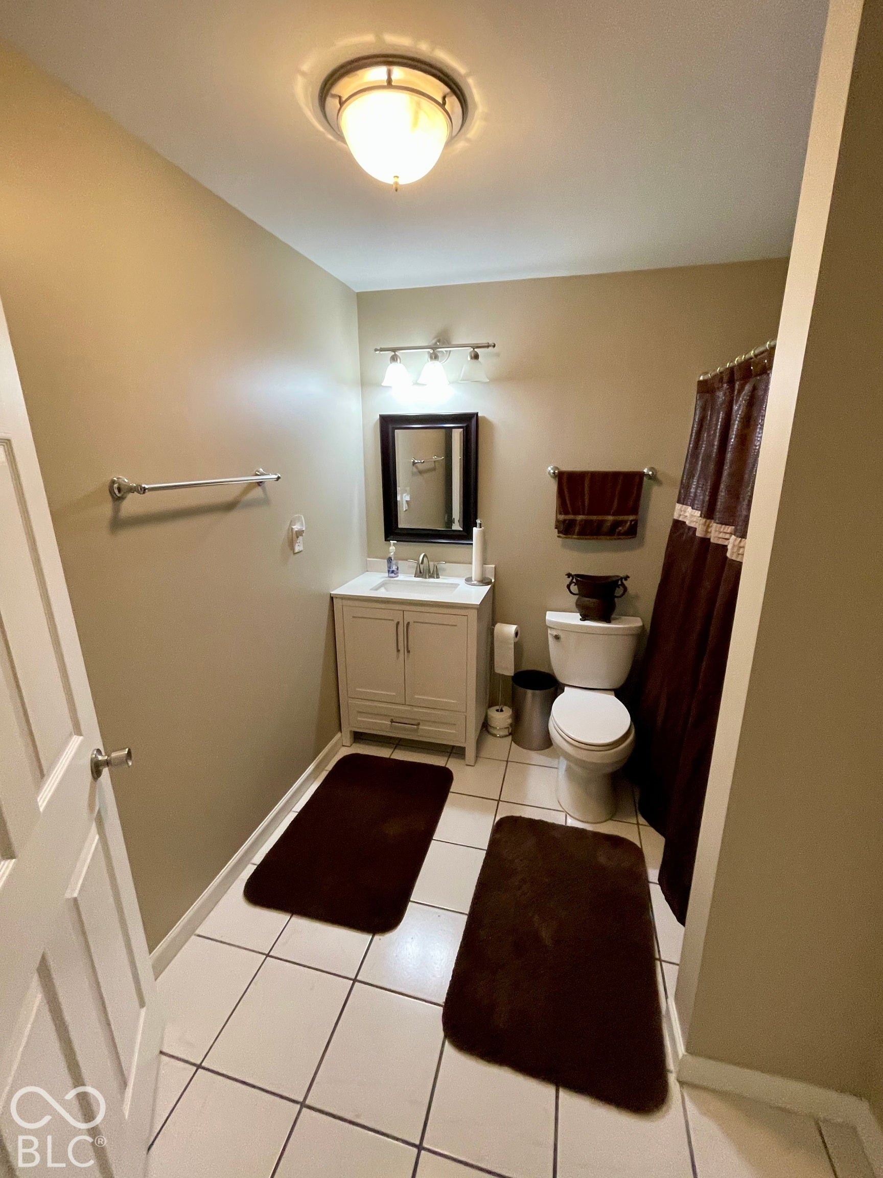 Recessed ceiling light fixture, white tile flooring, white vanity with cabinet storage, mirror with dark frame, and wall-mounted vanity lighting