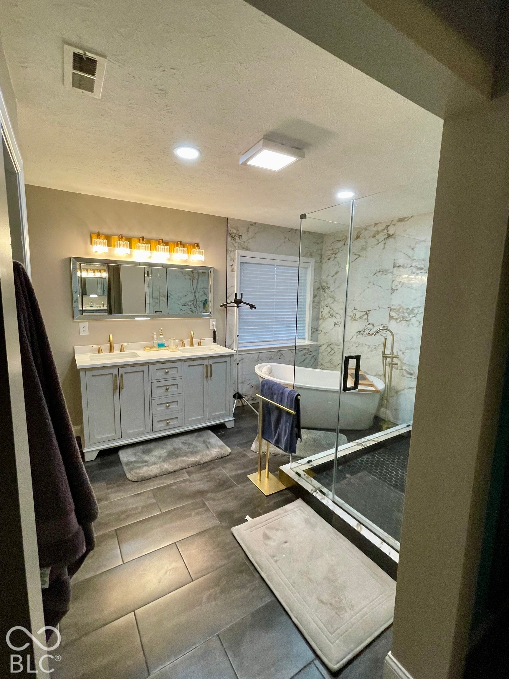 Spacious bathroom featuring a dual vanity with cabinet and white countertop, large format floor tiling, a freestanding soaking tub, a walk-in shower with clear glass enclosure, and recessed lighting