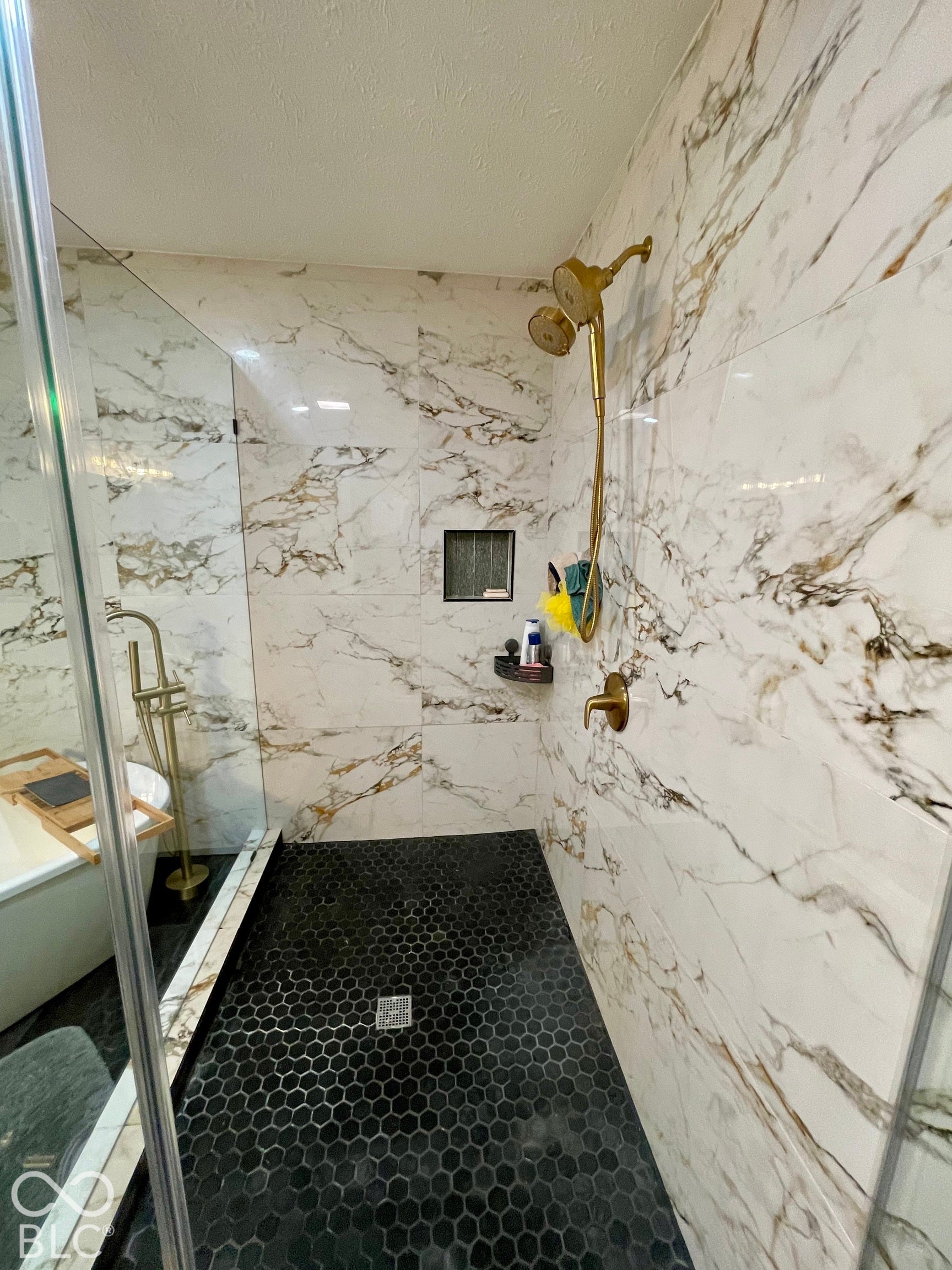 Walk-in shower featuring white and gold-veined wall tile, black hexagonal floor tile, and gold-finish fixtures