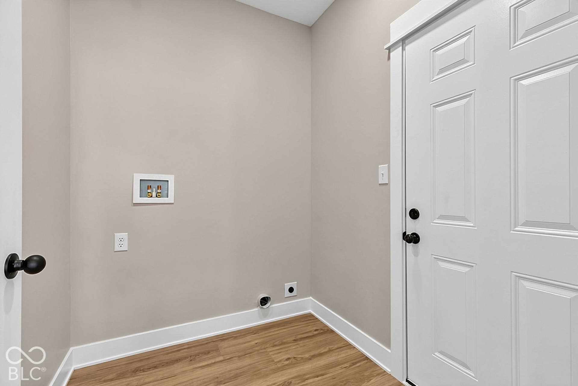Utility area featuring wood-finish flooring, a recessed water hookup box, and a white six-panel door with matte black hardware