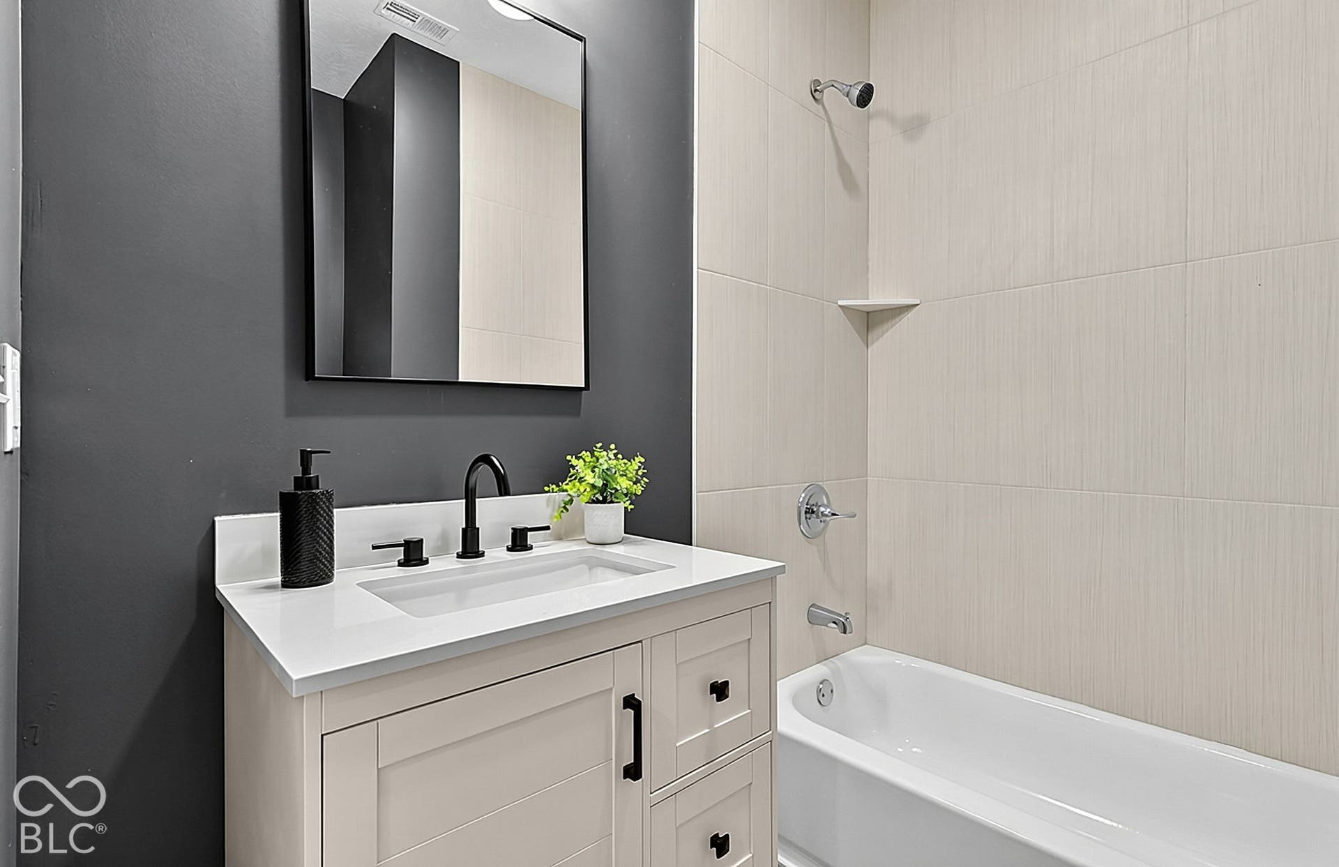 Bathroom featuring a vanity with a white countertop and rectangular undermount sink, black hardware, a framed mirror, and a tub with light-toned wall tile