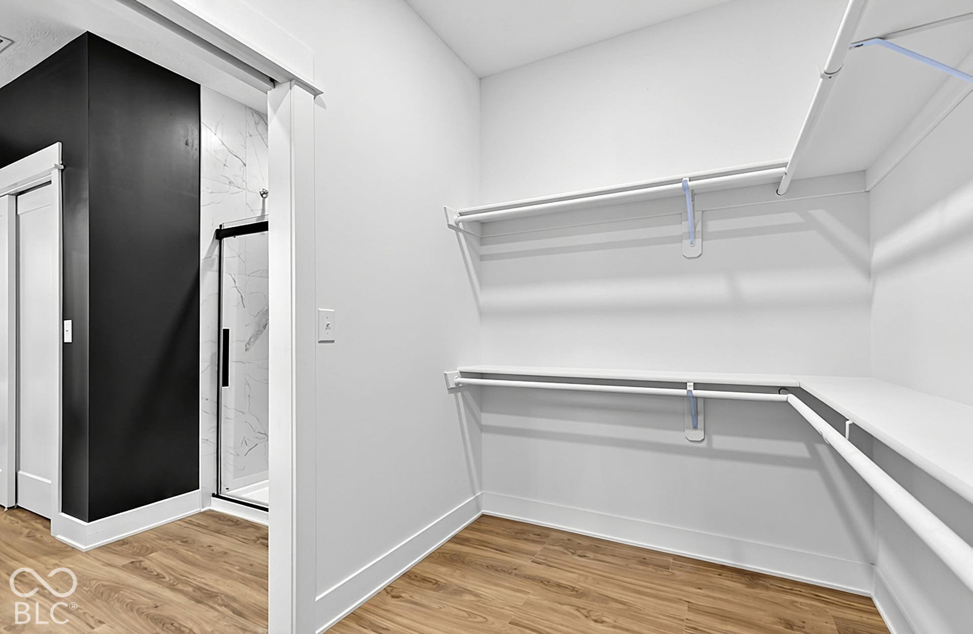 Walk-in closet featuring tiered white wire shelving, wood-finish flooring, and a doorway revealing a shower with a sliding glass door and stone-patterned tile surround