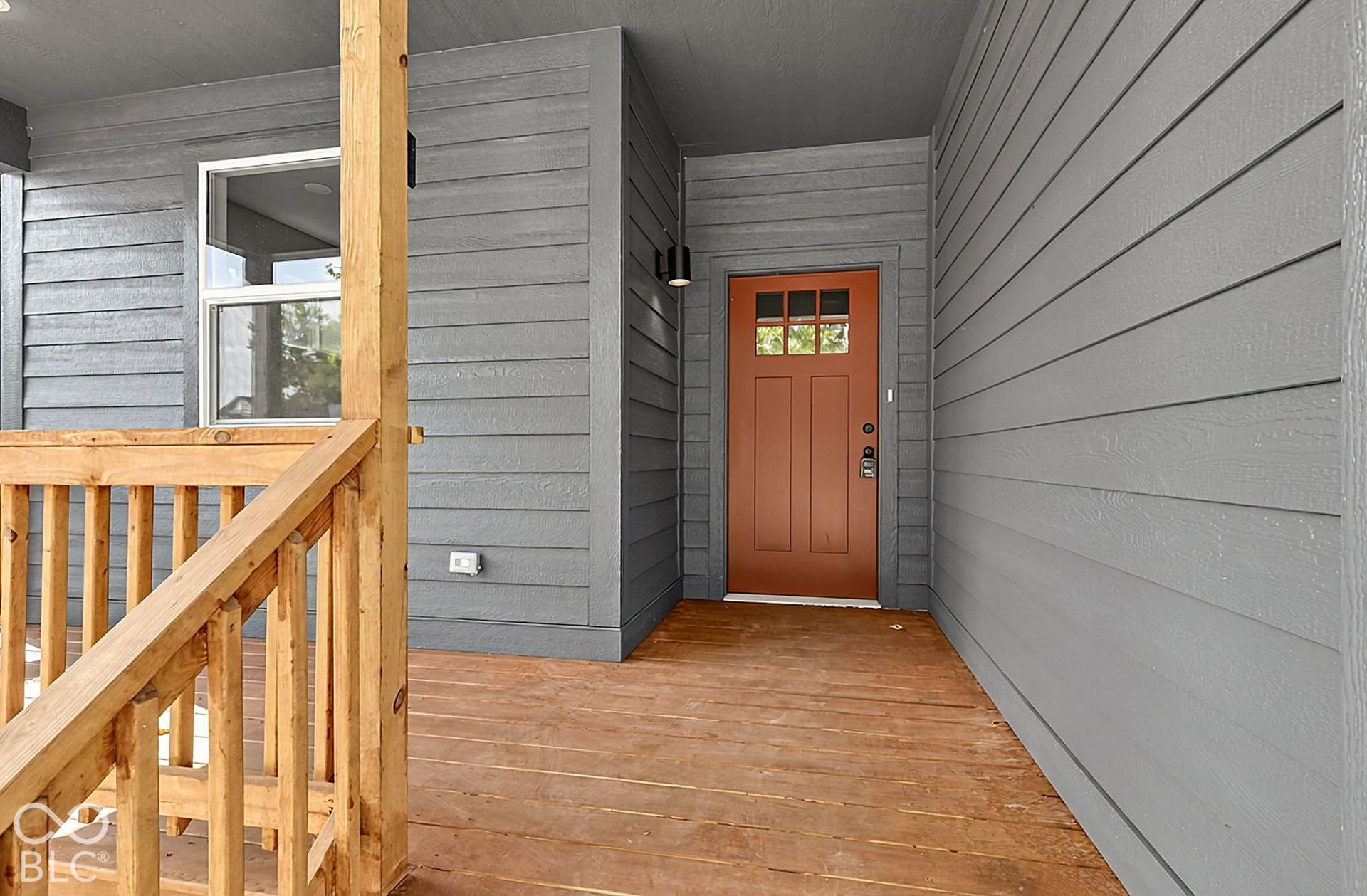 Covered front porch featuring wood-finish decking, horizontal lap siding, a rustic-style entry door with glass upper panels, and cylindrical wall-mounted exterior lighting