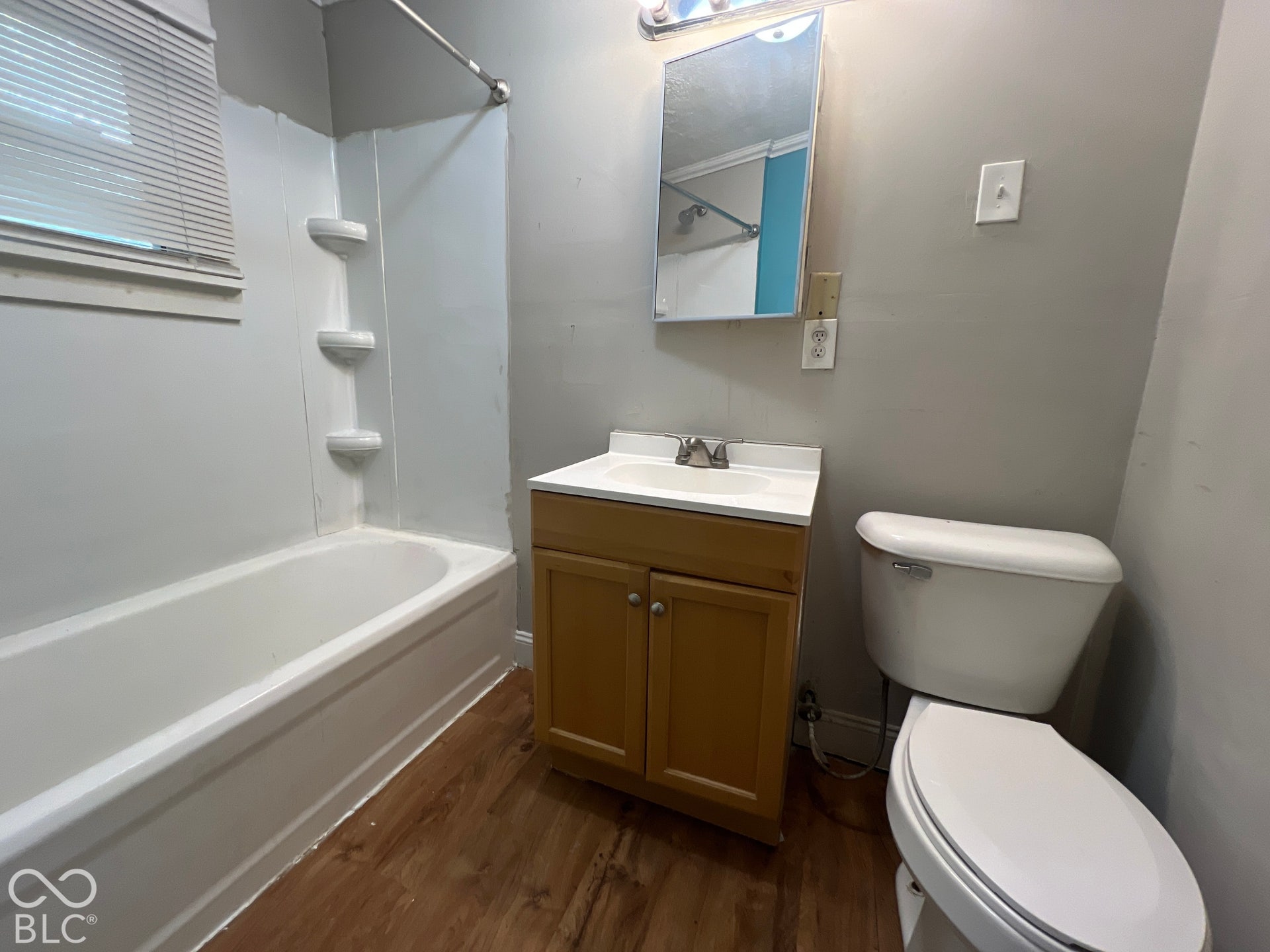 1431 W Pruitt Street, Indianapolis, IN 46208 Full bathroom featuring a wood-finish vanity cabinet with integrated sink and countertop, an enclosed shower-tub combination with white surround and chrome fixtures, ceramic toilet, medicine cabinet with mirror, wood-finish flooring, and neutral wall finishes