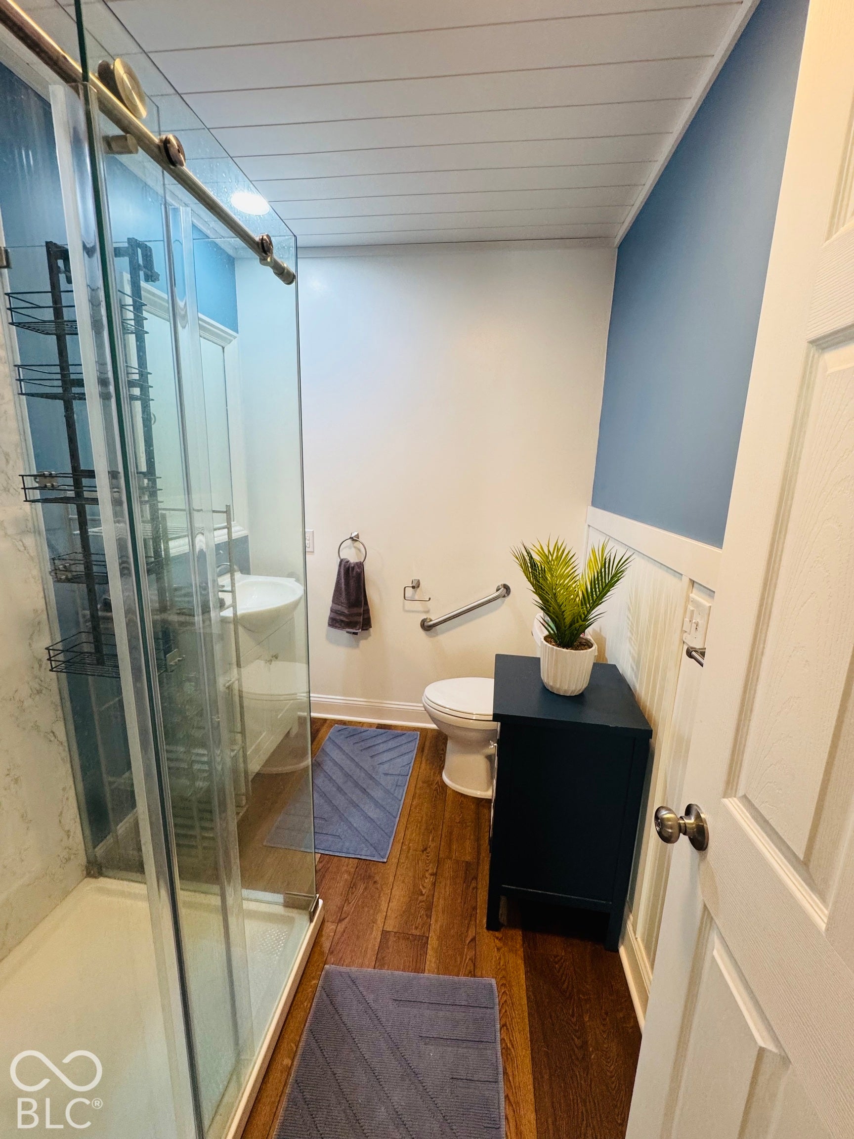 Glass-enclosed shower with sliding door, wood-finish flooring, white shiplap ceiling, and wainscoting