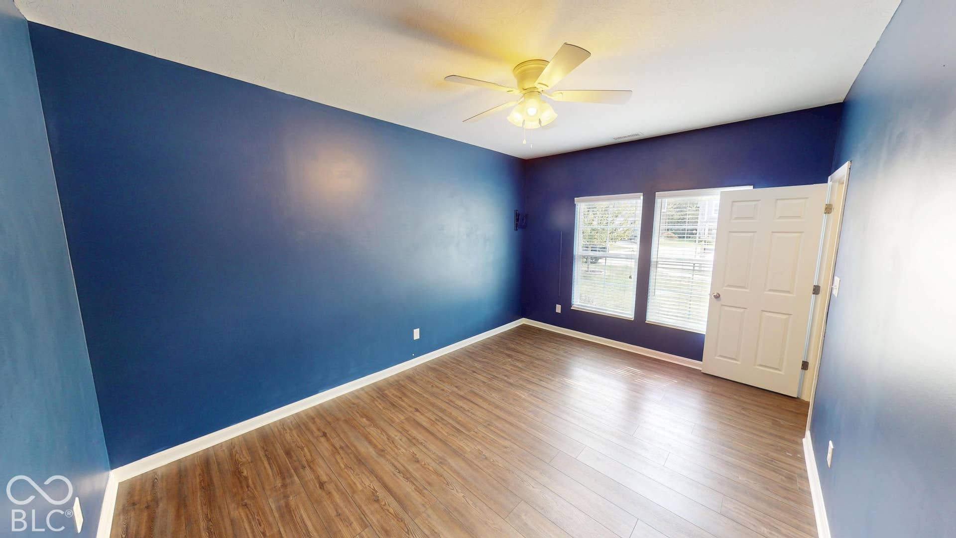 Wood-finish flooring, two large windows with blinds, a ceiling fan with integrated lighting, and a paneled door