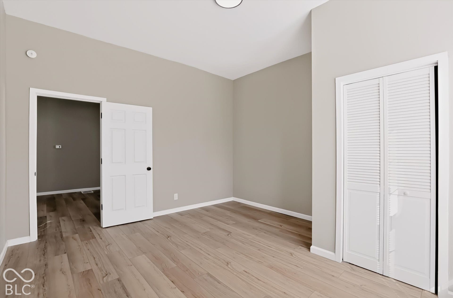 Neutral-toned interior featuring wood-finish flooring, high ceilings, and white paneled doors, including a closet with louvered double doors