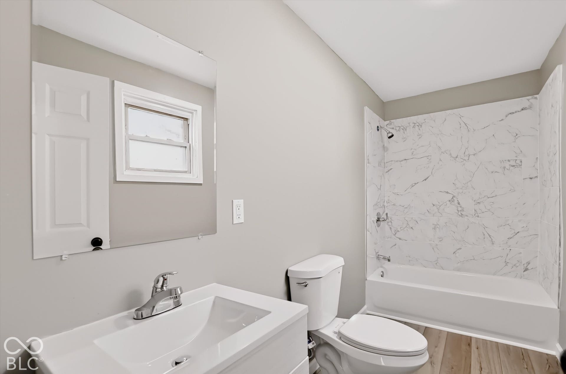 Bathroom featuring a white vanity with a rectangular basin, large wall-mounted mirror, wood-finish flooring, and a tub surround with a light-toned patterned finish