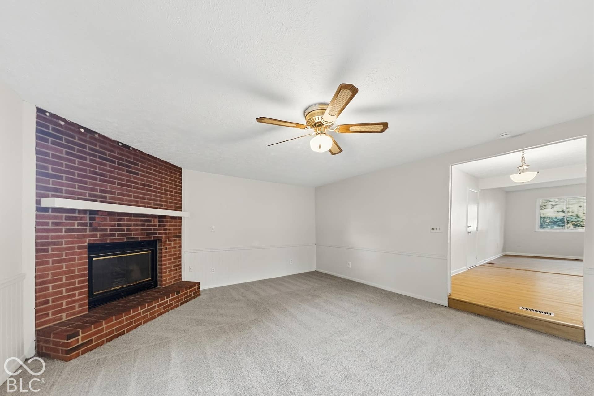 Brick fireplace with a white mantel, ceiling fan with integrated lighting, wall-to-wall carpeting, and white chair rail molding