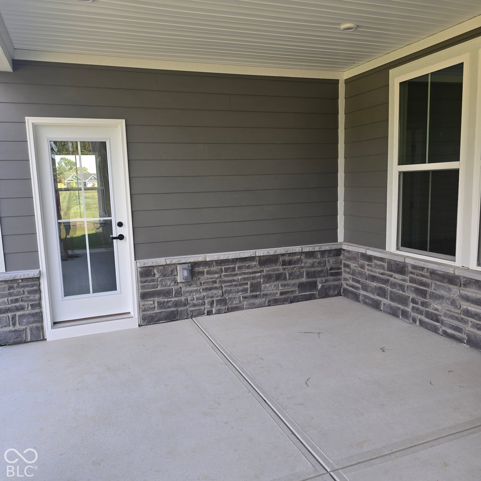 Covered patio featuring concrete flooring, a white-framed glass-paned door, gray siding, a stone accent wall, and a white-framed window