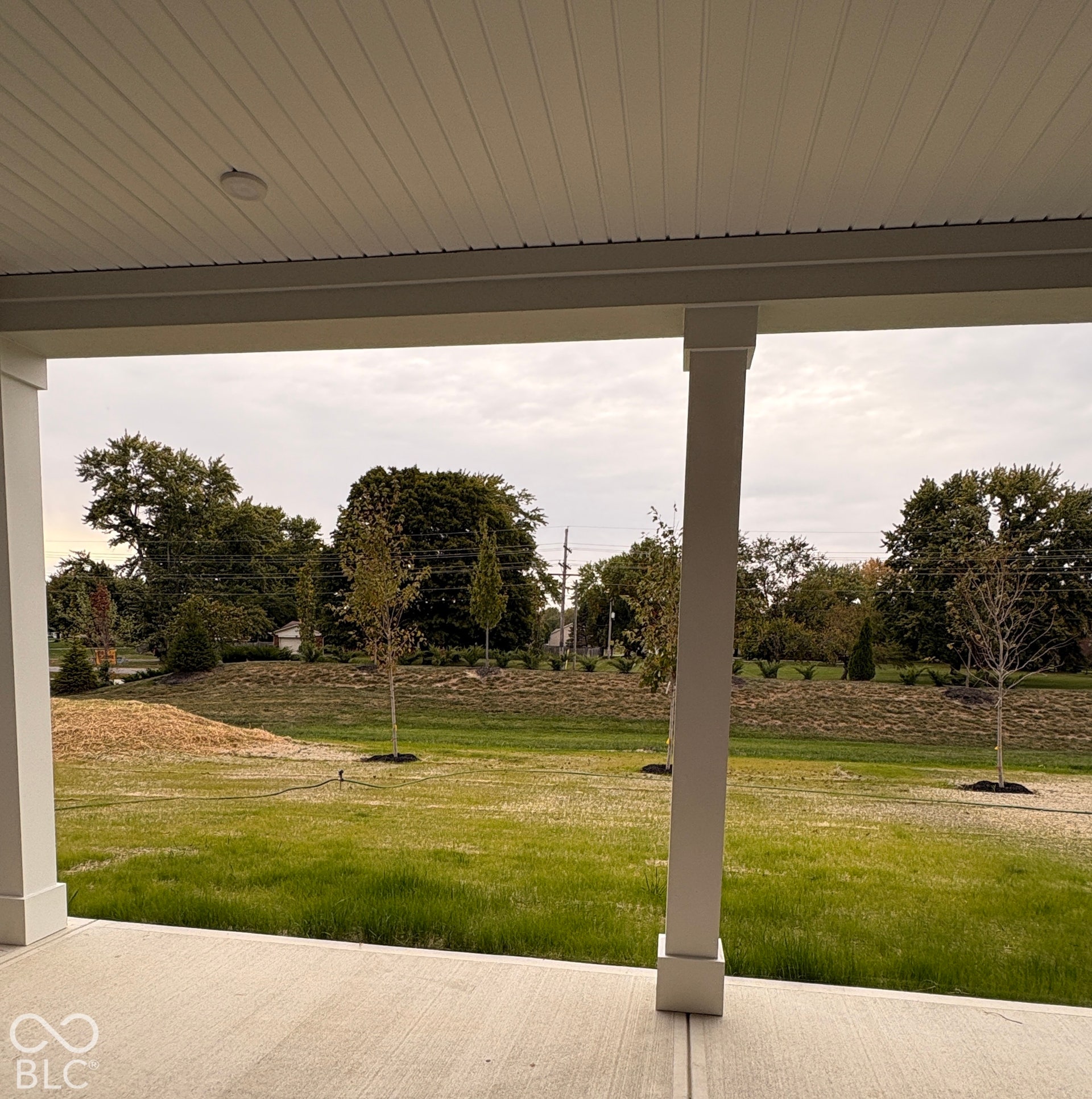 Covered concrete patio with a beadboard ceiling, recessed lighting, and a verdant grassy area with mature trees