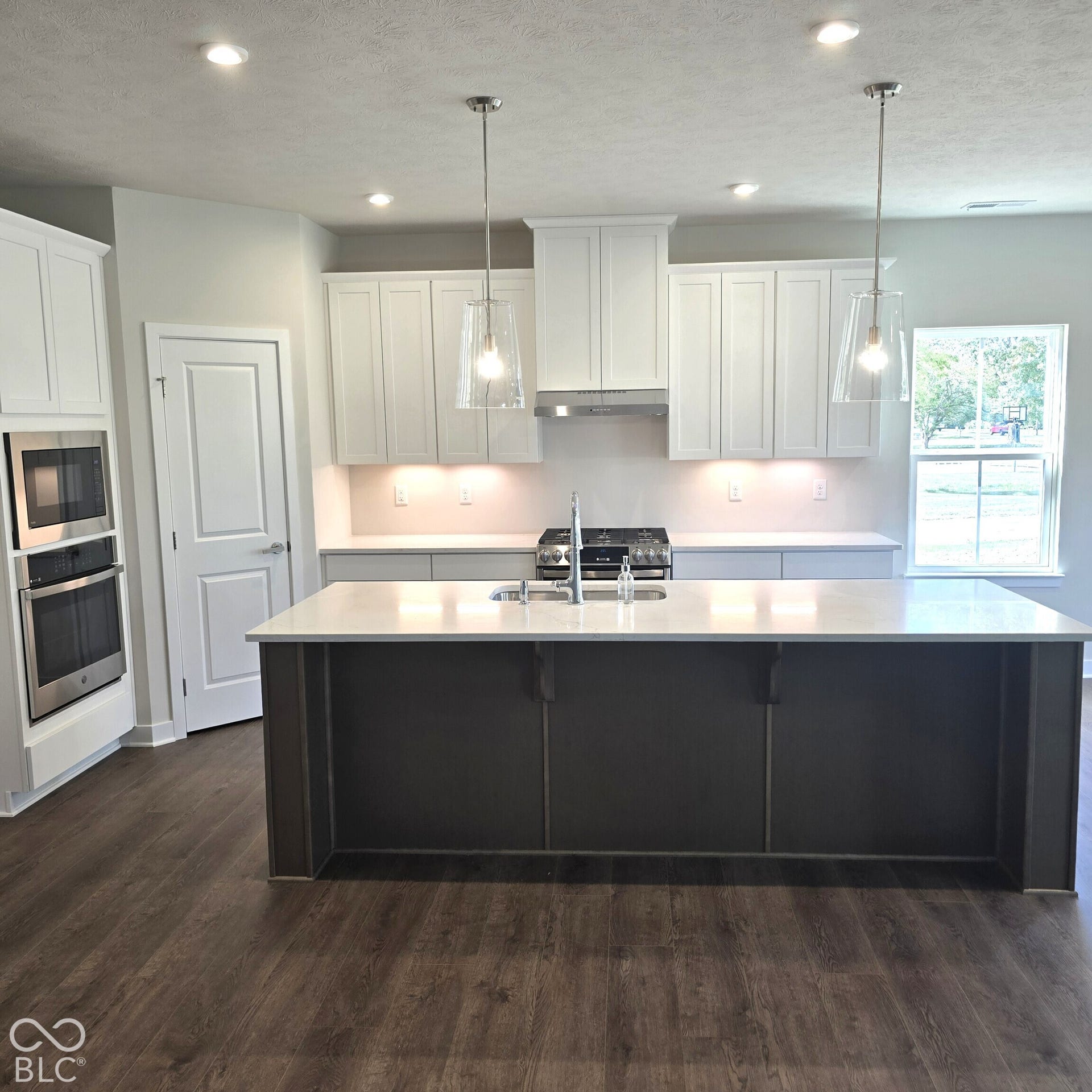 Expansive kitchen island with a built-in sink, white shaker cabinetry, stainless steel appliances, pendant lighting, and wood-finish flooring