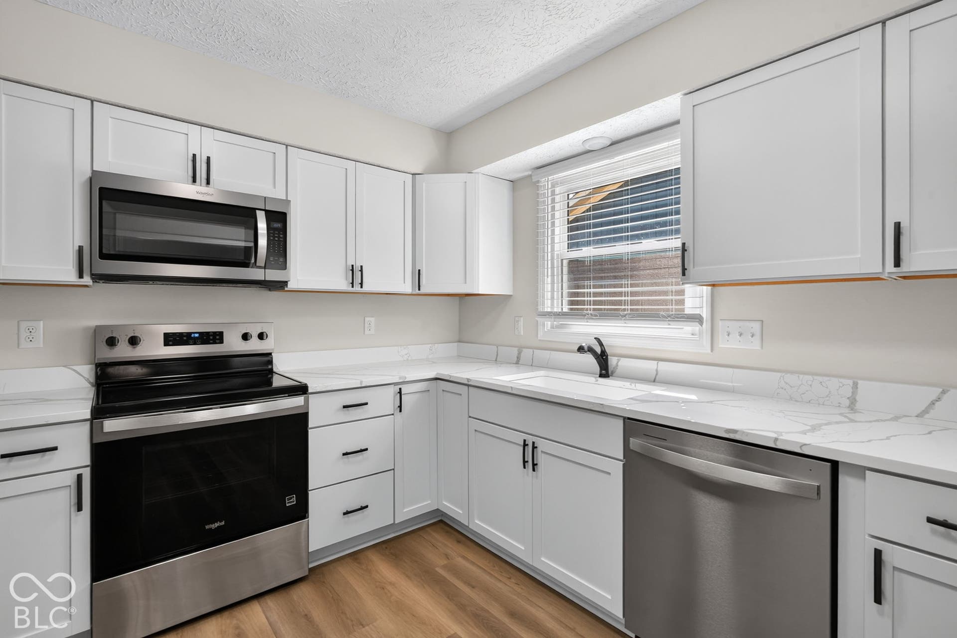White shaker-style cabinetry with matte black hardware, stainless steel appliances, light-toned countertops with veining, and wood-finish flooring