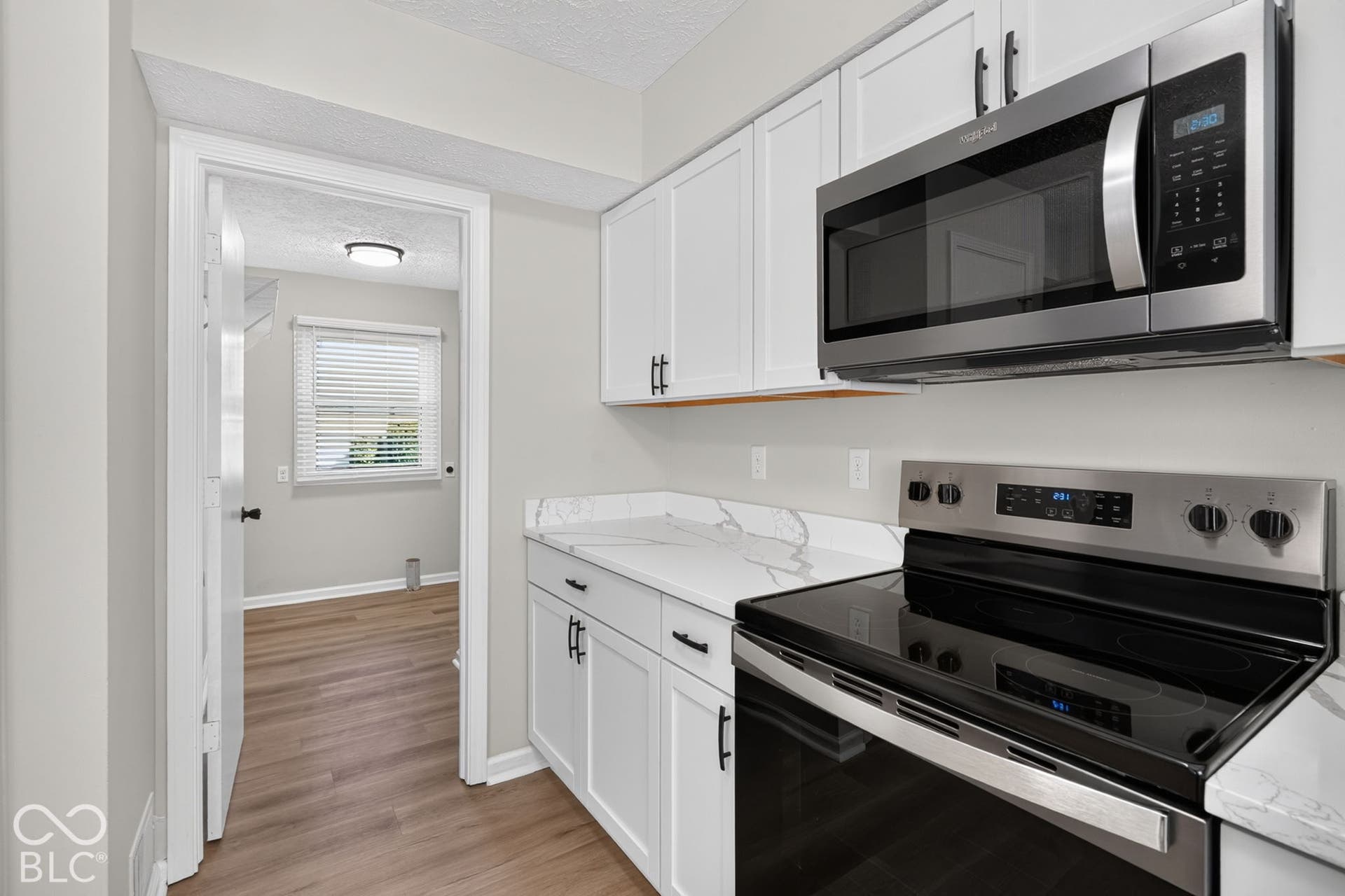 White shaker-style cabinetry, stainless steel microwave, black glass cooktop, wood-finish flooring, and white countertops with veining