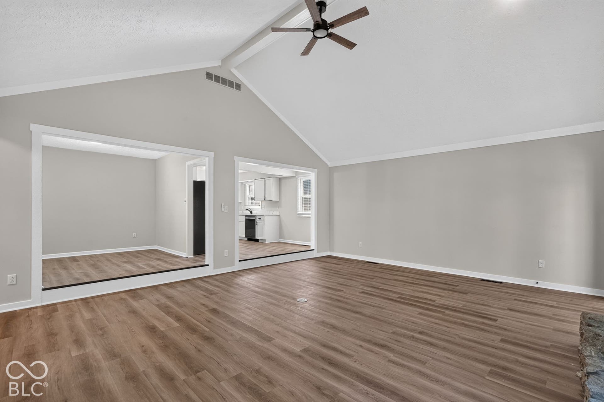 Vaulted ceiling, wood-finish flooring, ceiling fan, white trim, and a partial view of a kitchen with white cabinetry