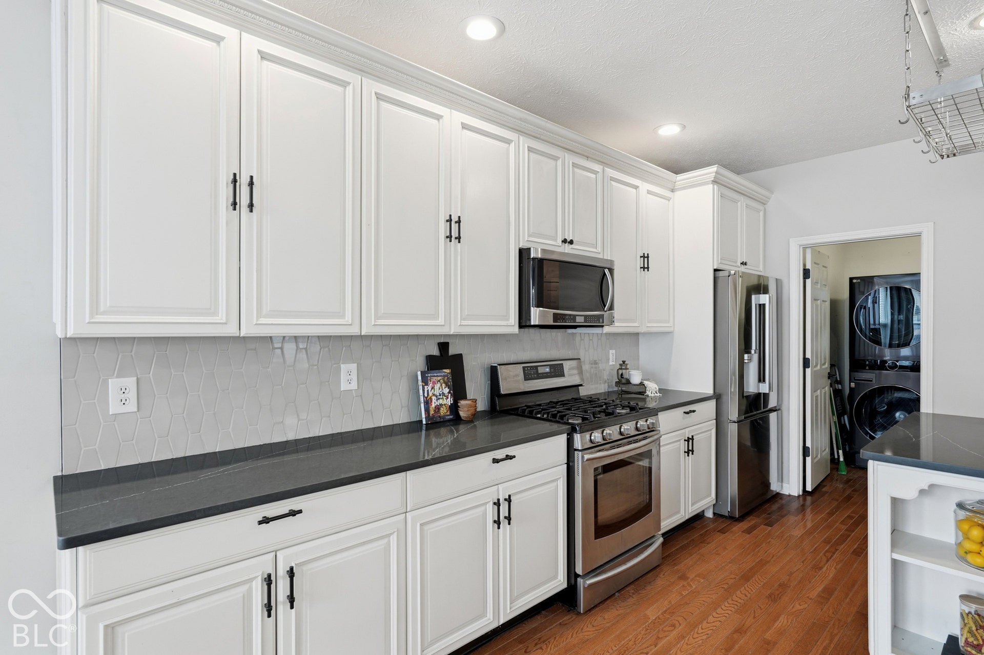 White shaker-style cabinetry with crown molding, stainless steel appliances, dark countertops, a geometric tile backsplash, and hardwood flooring