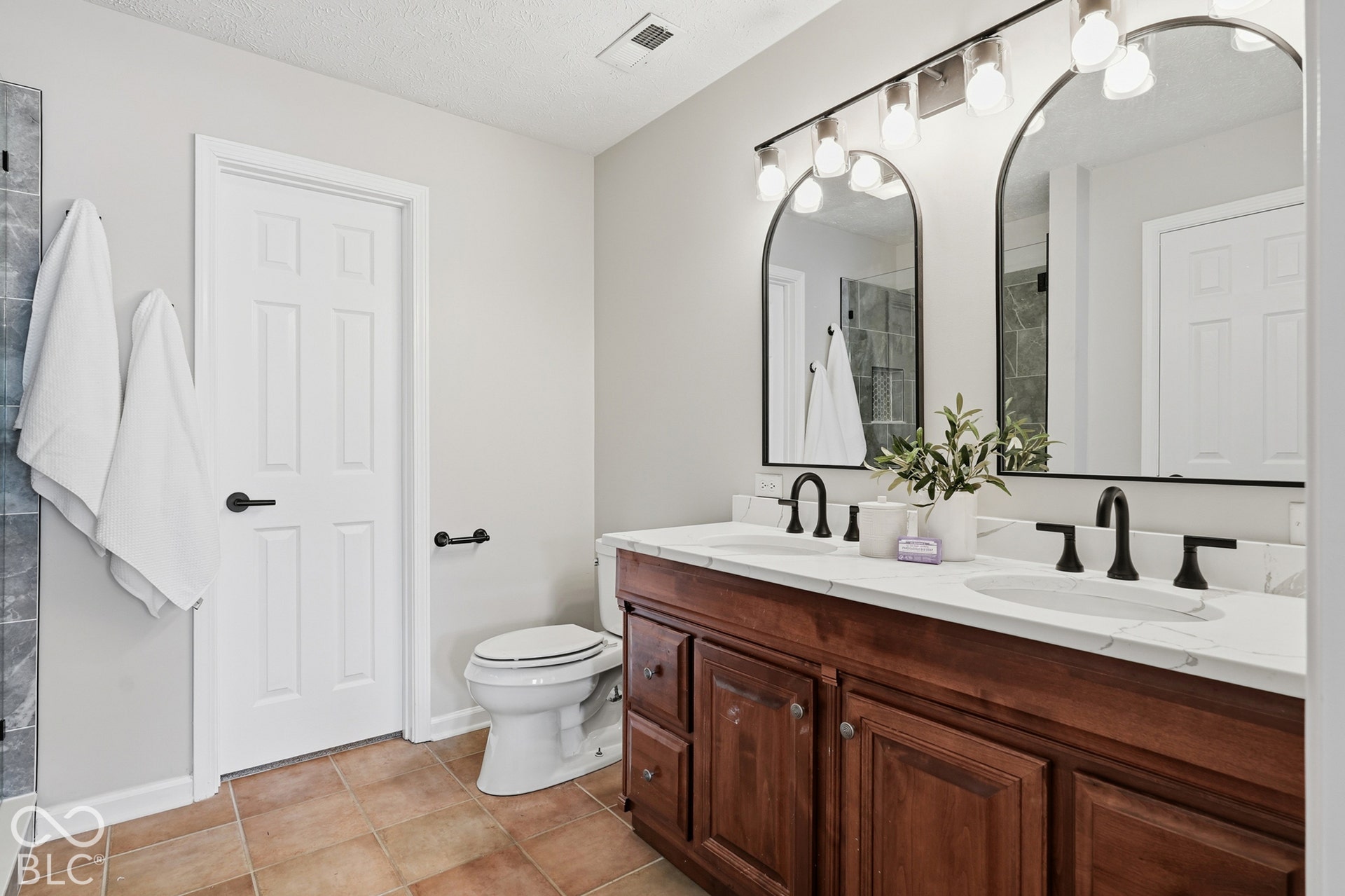 Dual arched mirrors with integrated lighting, a double vanity with wood-finish cabinetry, white countertop, and matte black fixtures