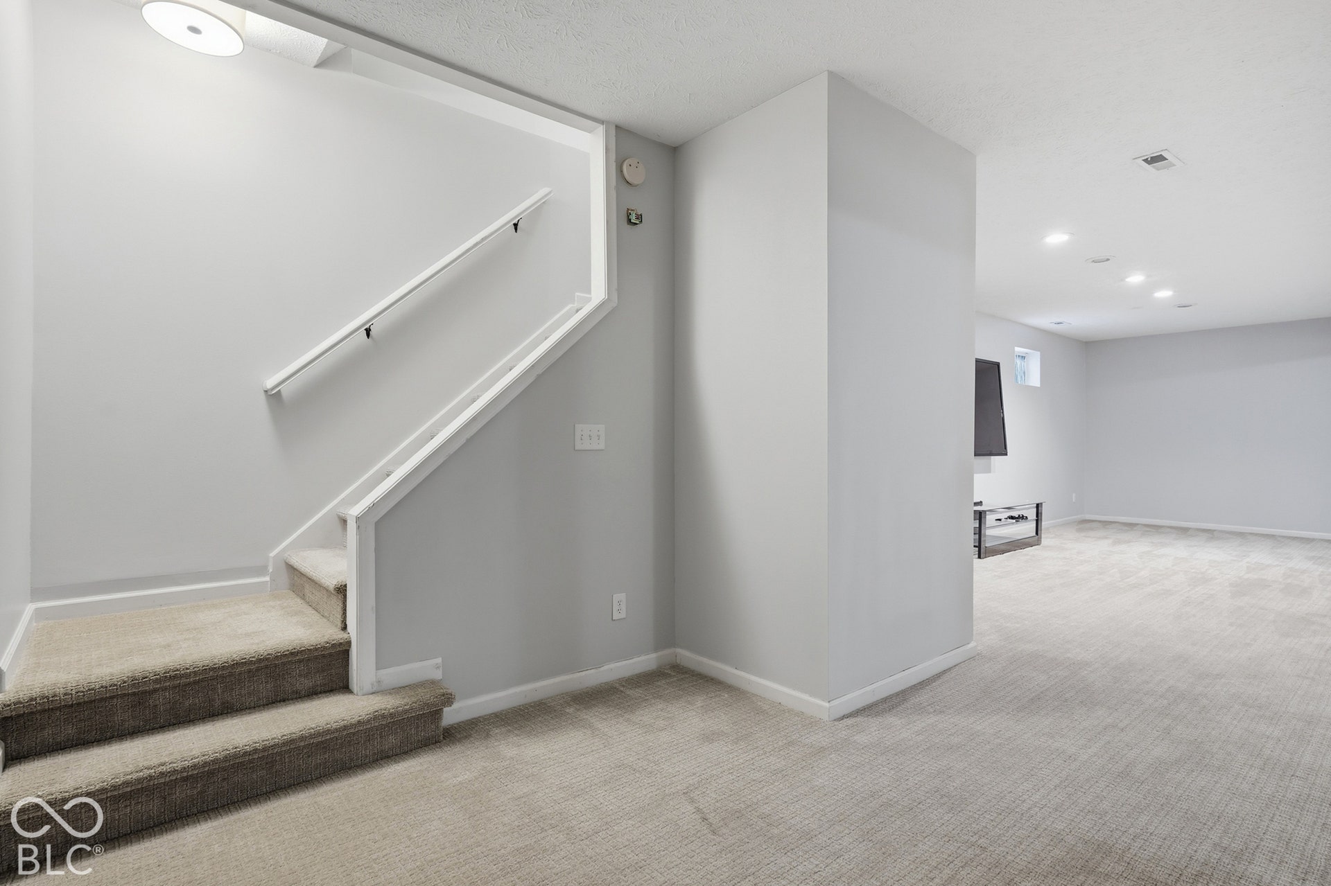 Carpeted staircase with white handrail leading to a spacious room with recessed lighting and neutral wall finishes